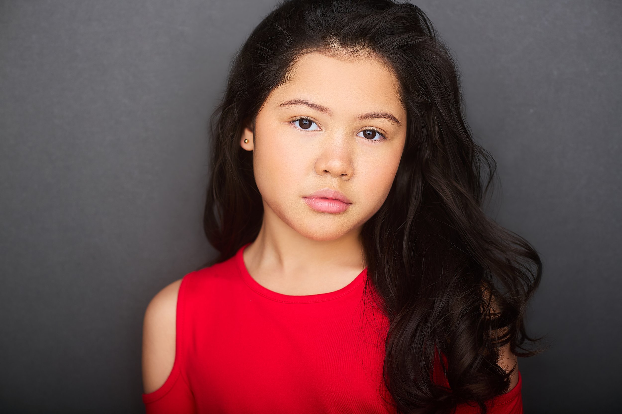 A young woman with long dark hair, wearing a red top with shoulder cutouts, poses against a gray background.