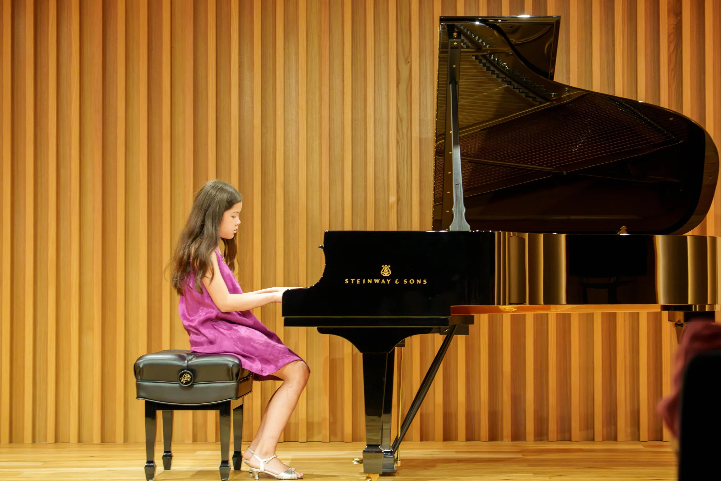 Young girl in a purple dress playing a black Steinway & Sons grand piano on a wooden stage with a wooden panel background.