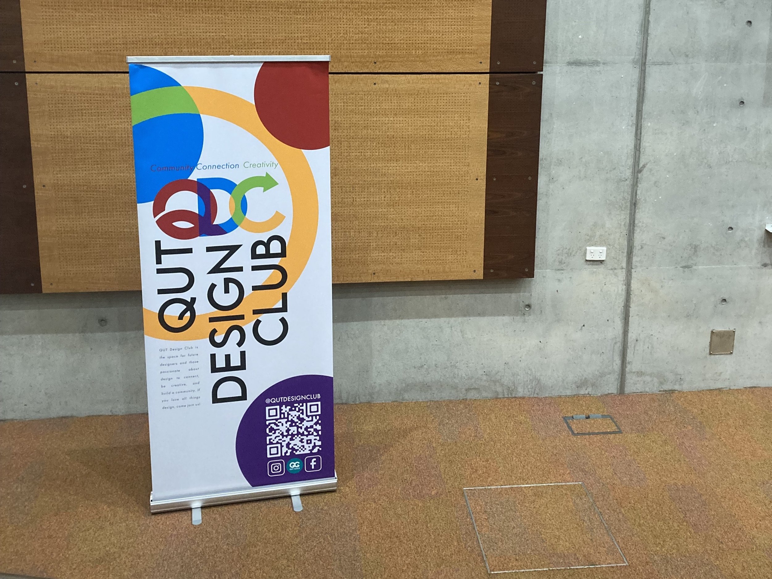 A vertical banner for QUT Design Club features colorful circles and the words 'Community, Connection, Creativity.'  The banner is placed indoors against a concrete wall.