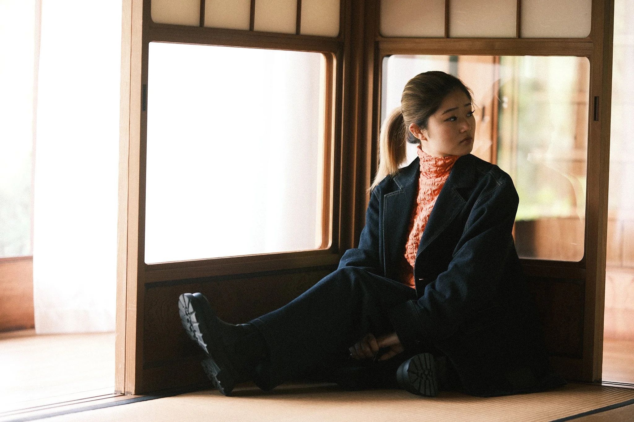Young woman sitting on the floor, leaning back against a wooden room divider, looking to her right with a contemplative expression, wearing a black coat, orange turtleneck, and black boots, inside a traditional Japanese room with tatami mats.