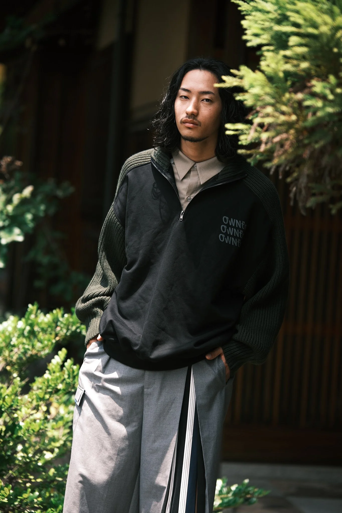 A young man with shoulder-length black hair and a goatee stands outdoors, wearing a black and green jacket over a beige collared shirt, with hands in his pockets, surrounded by greenery and a wooden structure.