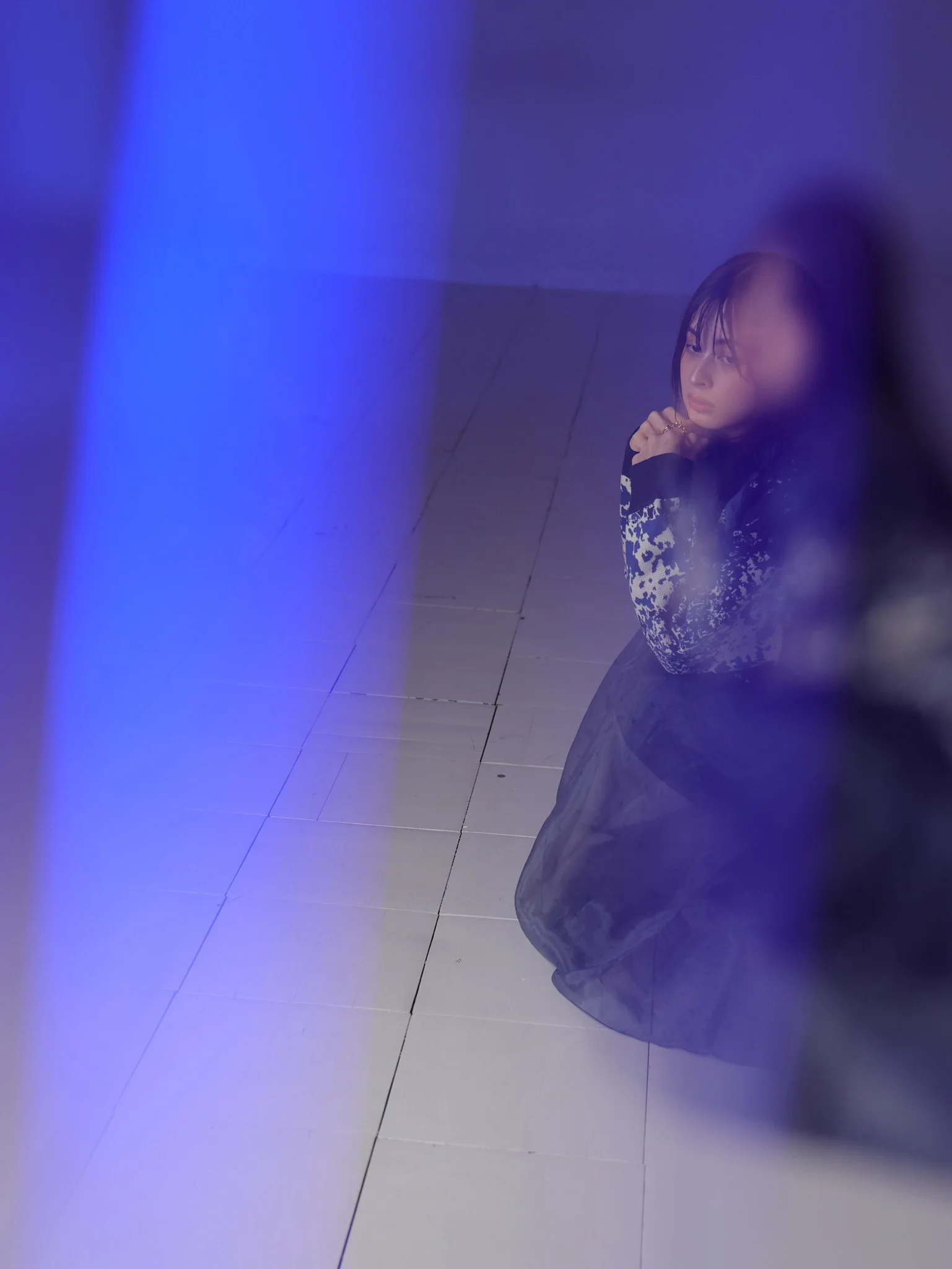 A young woman kneeling on the tiled floor, viewed through a blue-tinted transparent surface, with her hands clasped near her face and looking contemplative.