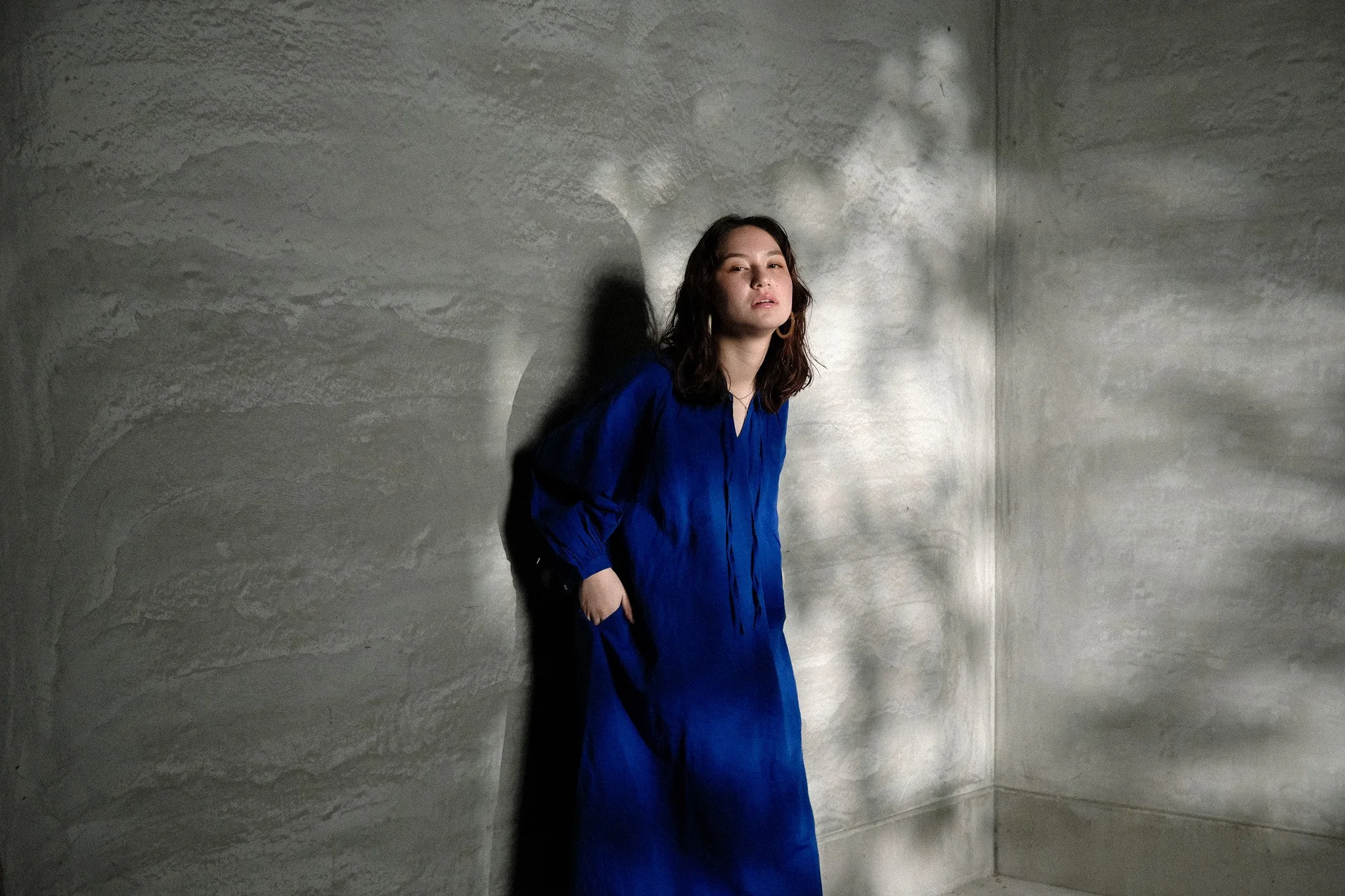 Woman in a blue dress leaning against a textured wall with light casting shadows behind her.
