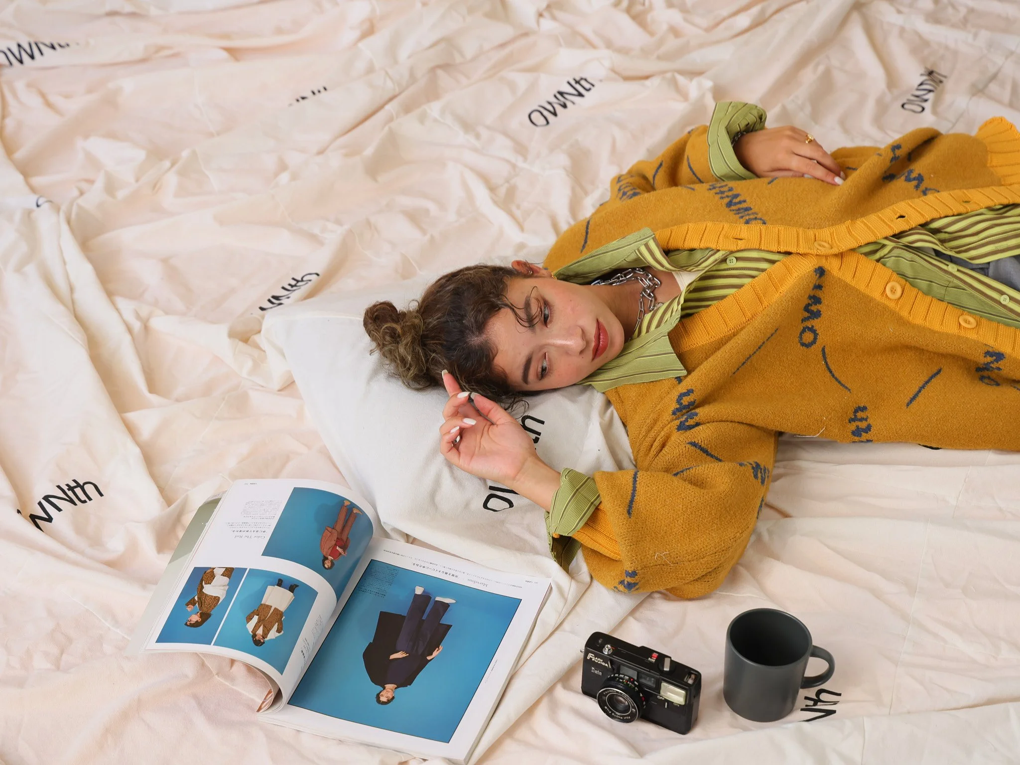 Young woman lying on her side on a bed, looking at an open magazine. She has curly hair, a chain necklace, and is dressed in a green striped shirt layered with an orange cardigan with blue writing. There are a camera and a dark gray mug next to her o