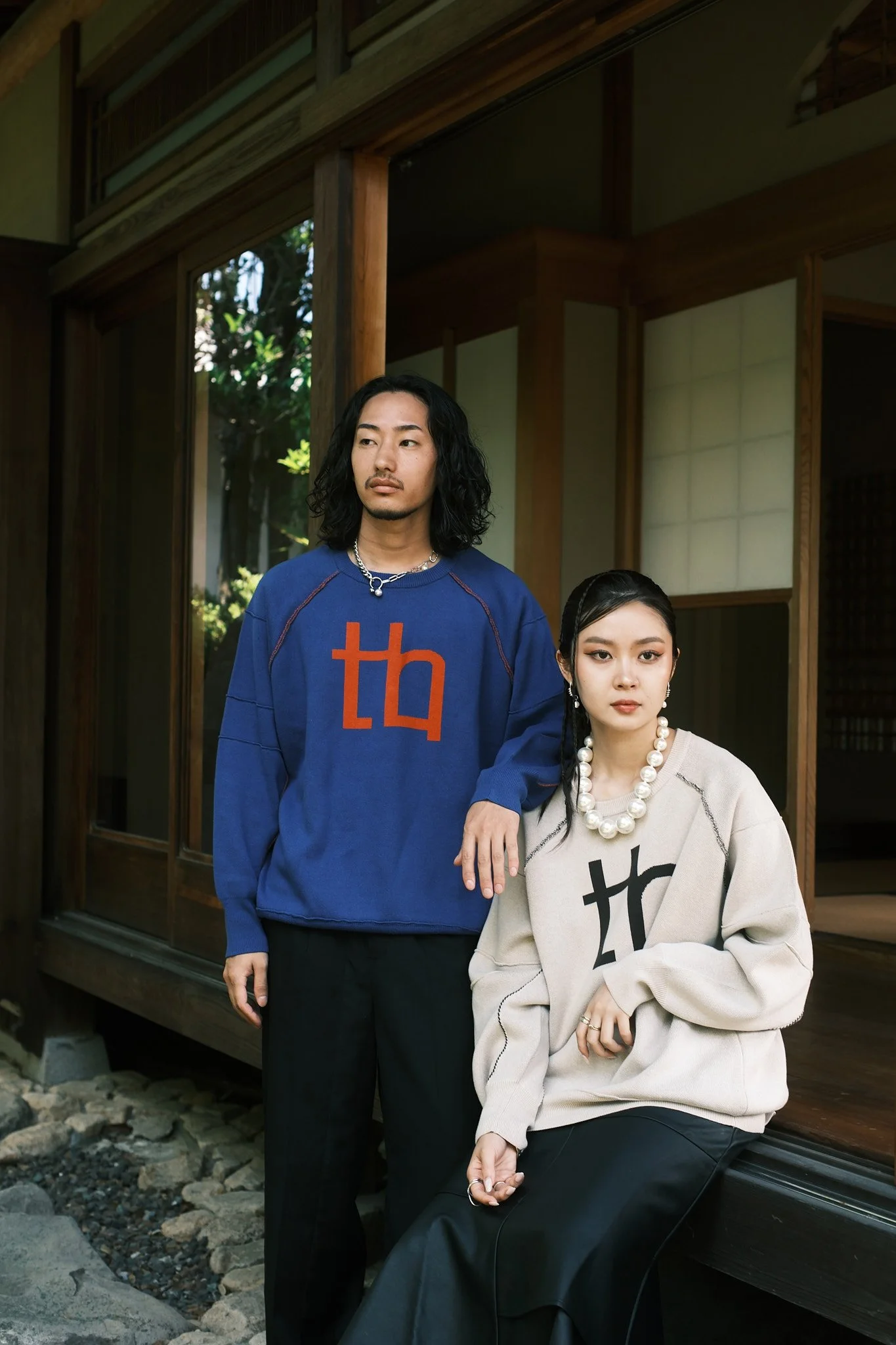 A man and woman pose outside a traditional Japanese house. The man is standing, wearing a blue sweatshirt with a red logo, and the woman is sitting, wearing an oversized cream-colored sweatshirt with a black logo and a large pearl necklace.