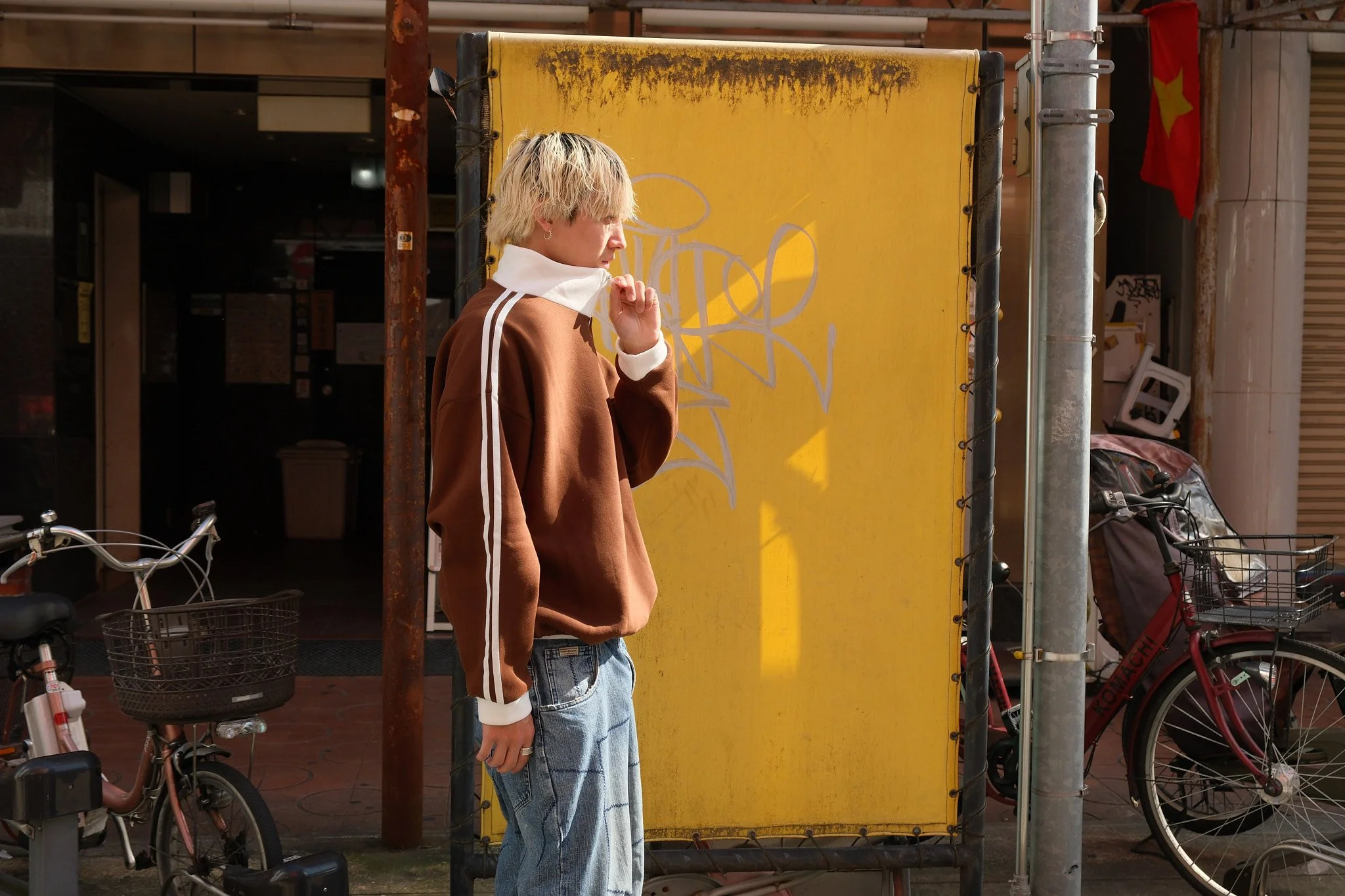 A person with blonde hair wearing a brown hoodie with white stripes on the sleeves and light blue jeans, standing on a city sidewalk near two parked bicycles, with a yellow signboard and a red flag in the background.