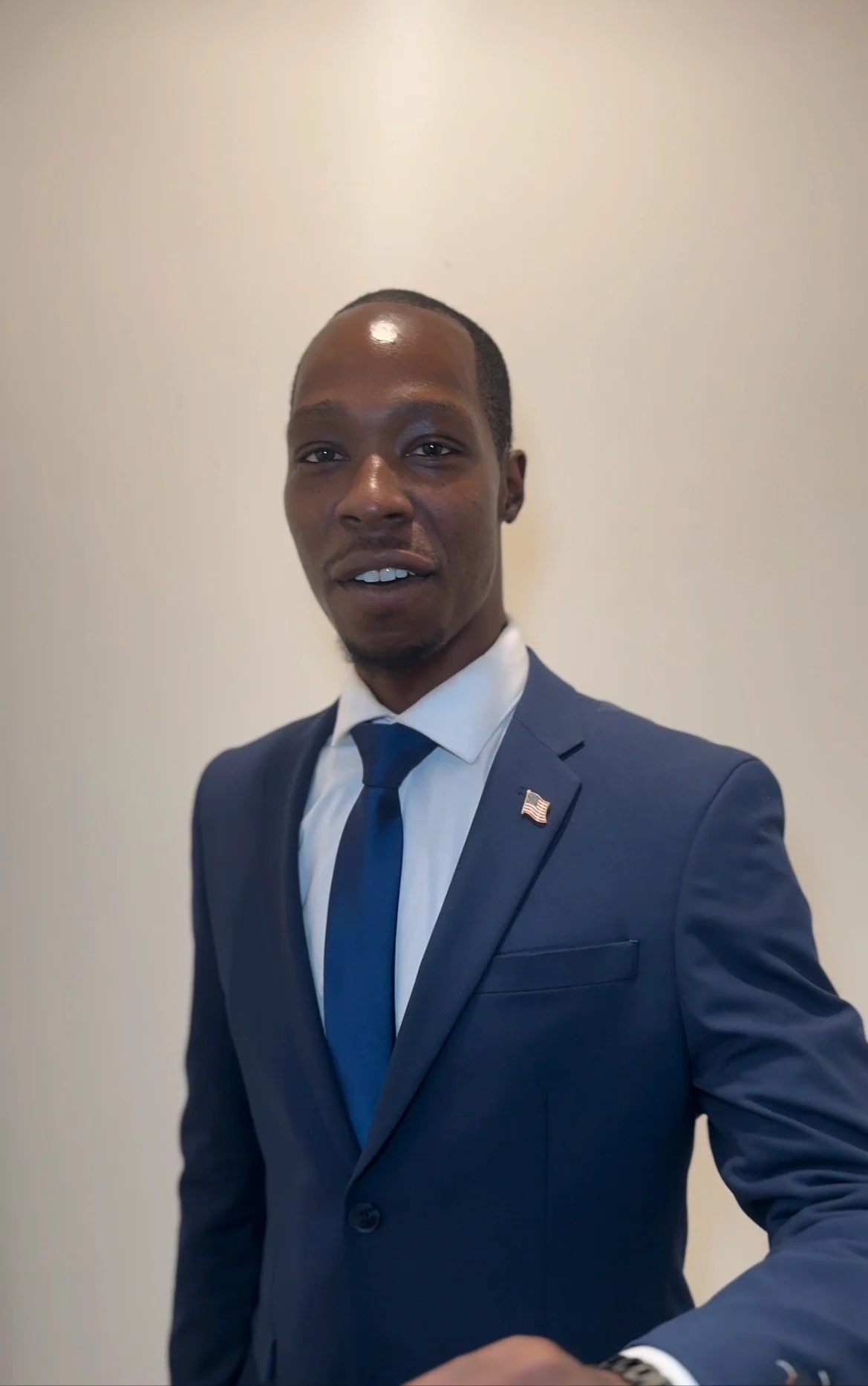 A man in a navy blue suit, white shirt, and blue tie, wearing a small American flag pin on his lapel, standing against a plain wall.