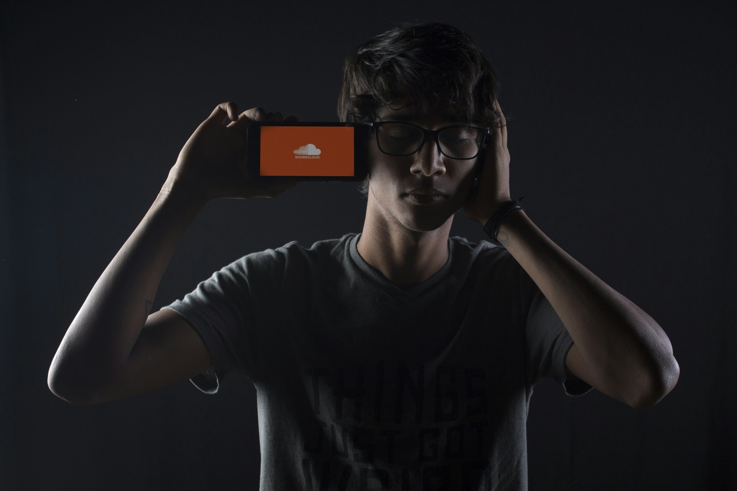 Young man with glasses holding a phone to his head displaying the SoundCloud logo, in a dark room. Soundcloud Link
