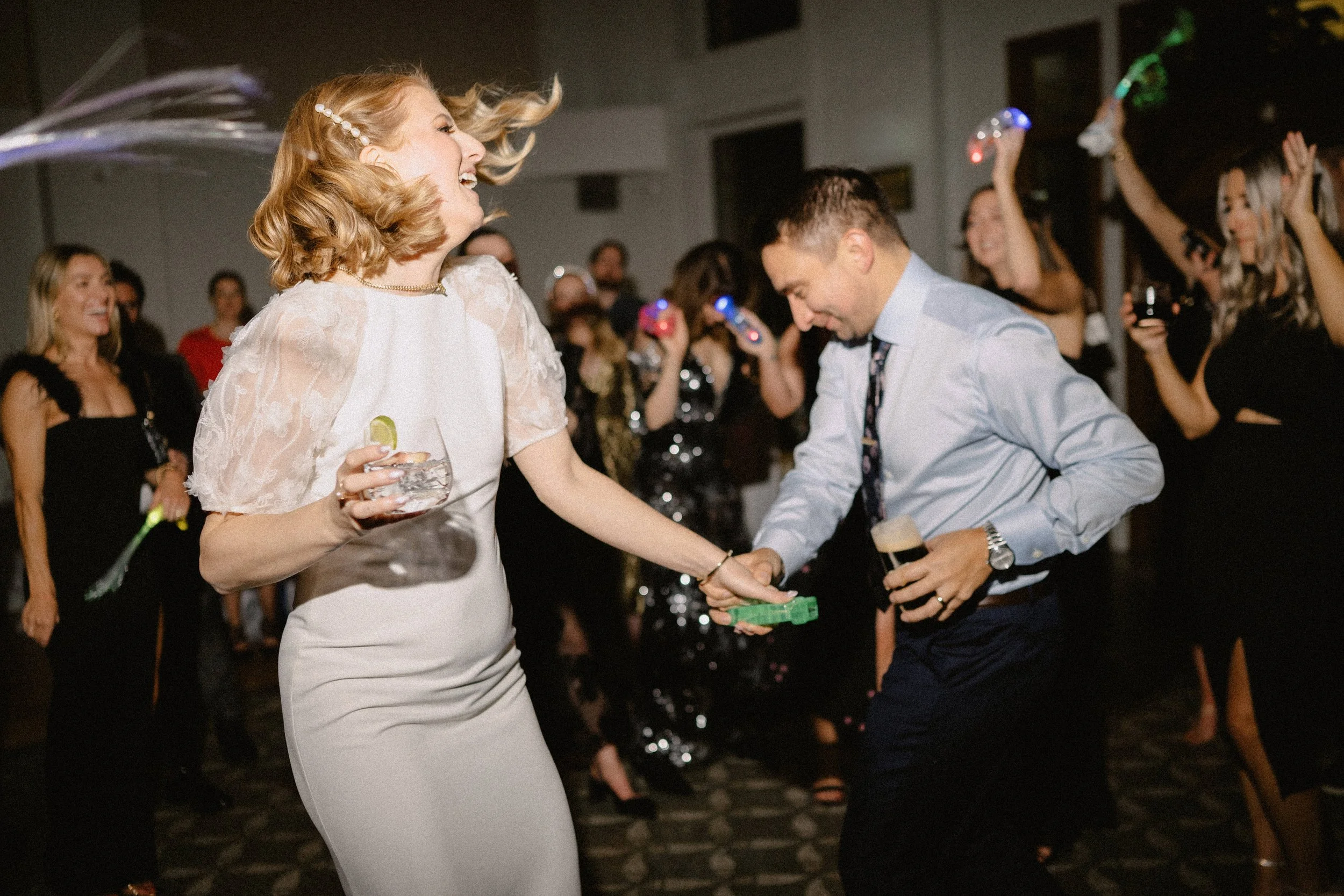People dancing and celebrating at a party or wedding reception, with a woman holding a drink and a man in a white shirt and tie holding a beer, surrounded by others with drinks and party accessories. Wedding Celebration. Wedding Dancefloor DJ