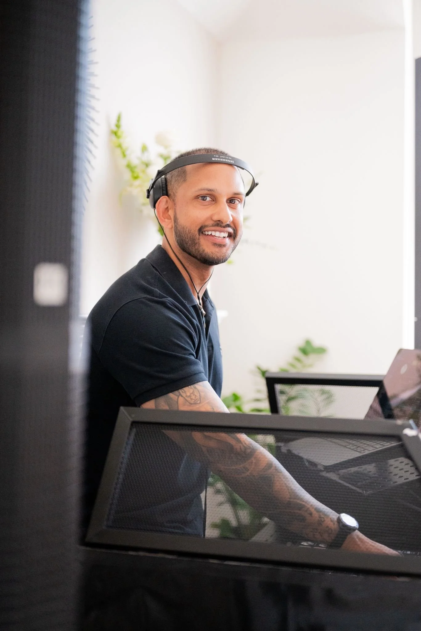 A smiling DJ wearing a black polo shirt and headphones sitting at a desk with a laptop and a view of a bright room with white walls and plants. DJing a private event in Vancouver.