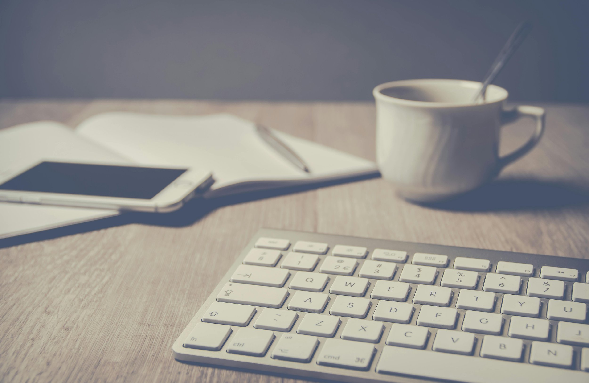 A white computer keyboard on a wooden desk, with a notebook, pen, smartphone, and a coffee mug with a spoon inside.