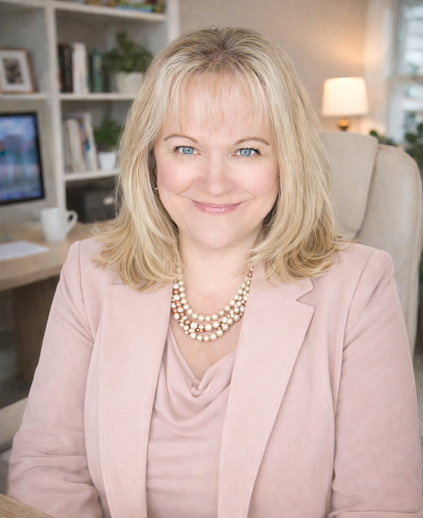 Portrait of a woman with blonde hair, blue eyes, wearing a pink blazer and pearl necklace, smiling at the camera in a home office setting.