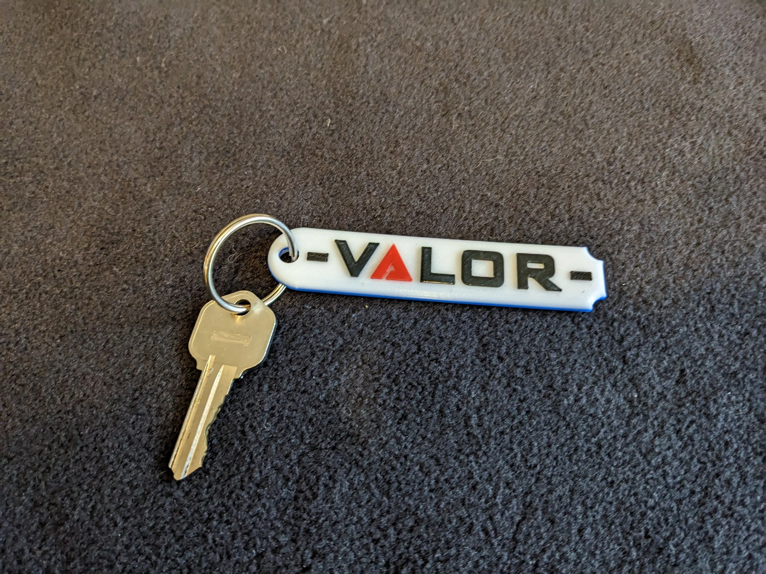 A roughly rectangular white keytag rounded on the left side and scalloped on the right, bearing the Alliance RV model name "Valor" in black lettering matching the company font and graphics.  Pictured with a gold key attached via a silver keyring