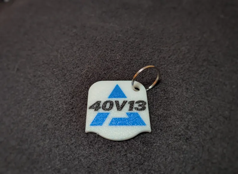 A white plastic keytag, roughly square in shape with rounded corners and a scalloped bottom bearing a blue Alliance company stylized "A"  in blue, and black lettering integrated, displaying the Alliance RV model name "40v13"