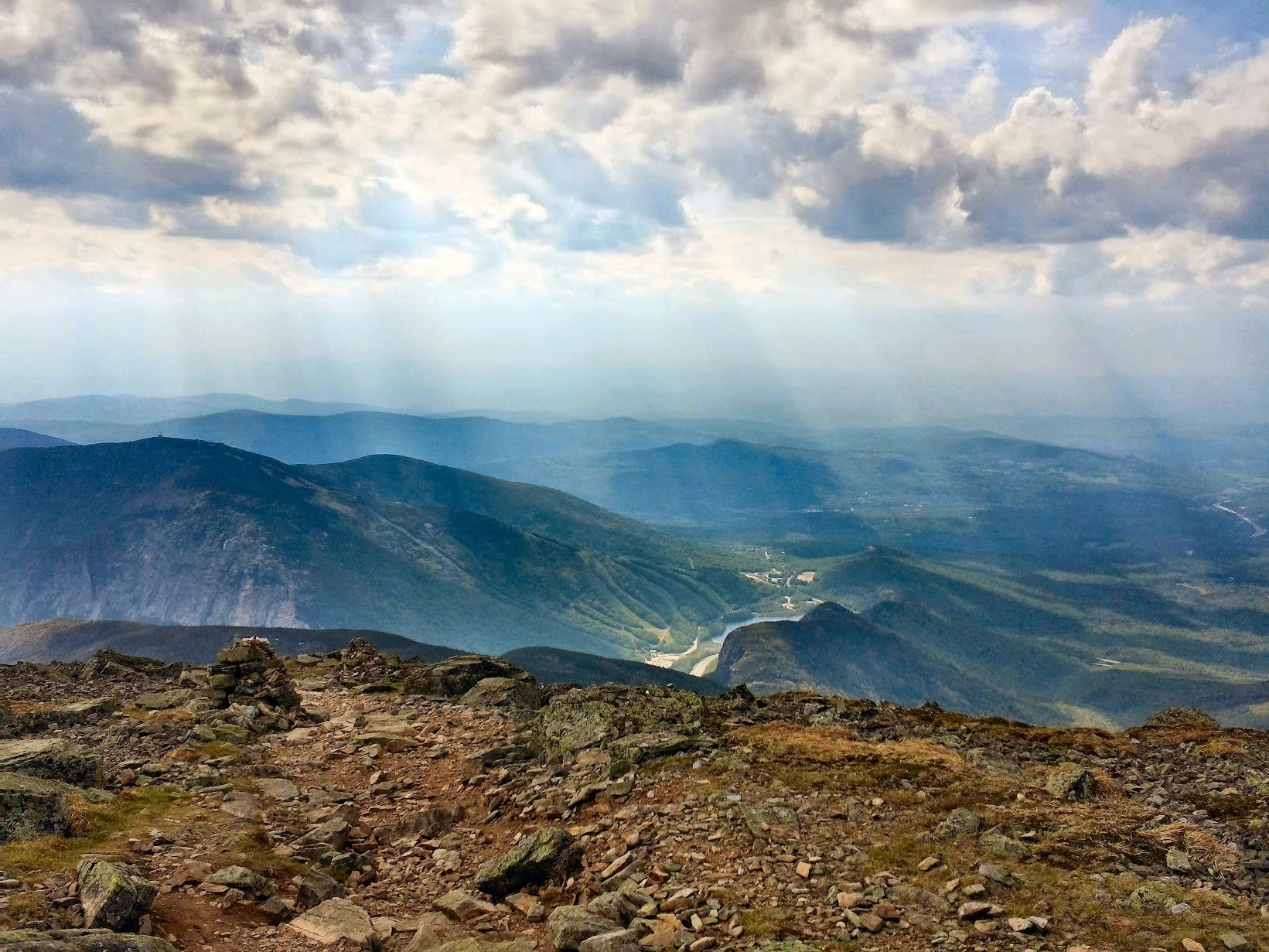 View from atop a mountain with a rocky dirt path in the foreground, overlooking green mountain ranges and valleys under a partly cloudy sky.