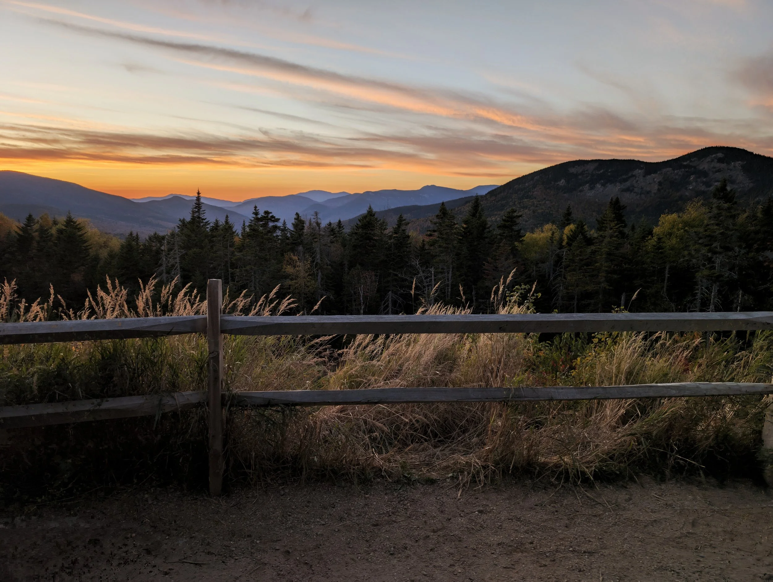 Sunset over mountain range with forest and grassy foreground, wooden fence in the front.