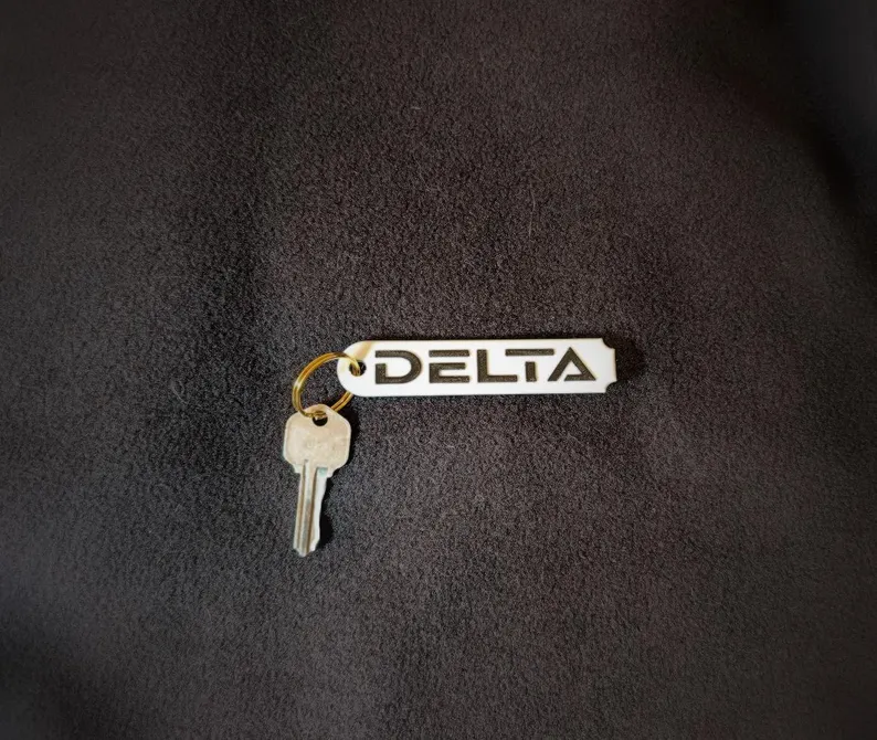 A roughly rectangular white keytag rounded on the left side and scalloped on the right, bearing the Alliance RV model name "Delta" in black lettering matching the company font and graphics.  Pictured with a gold key attached via a gold keyring