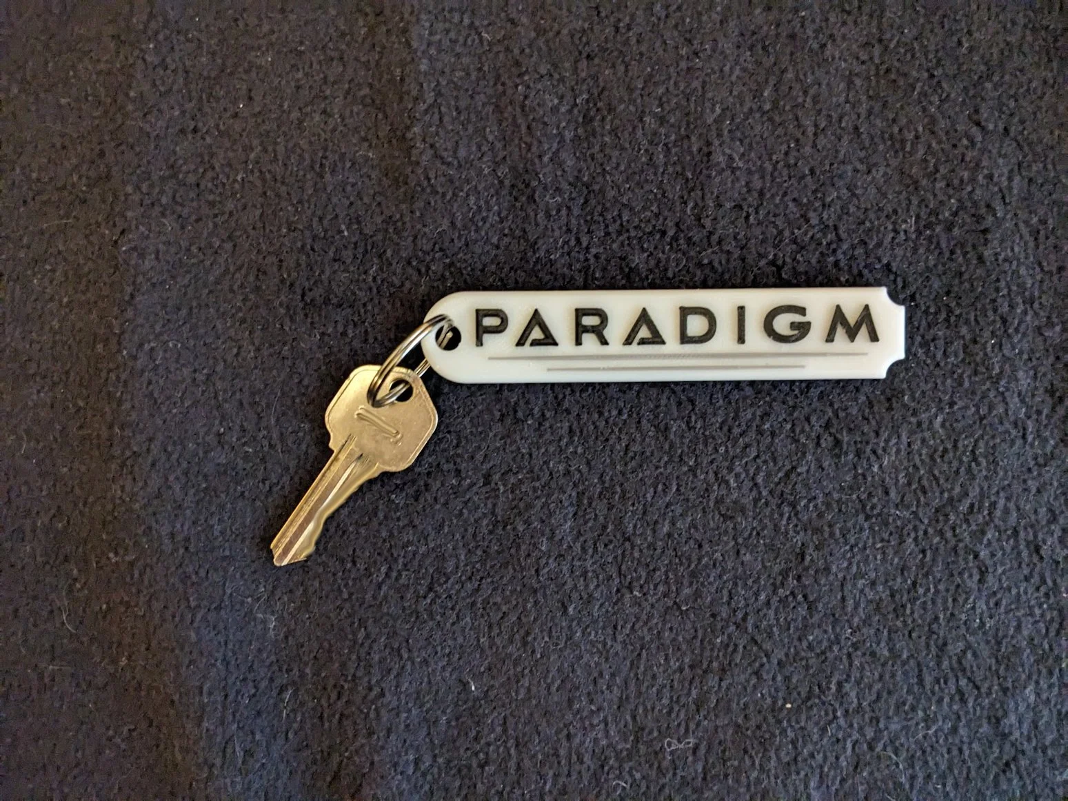 A roughly rectangular white keytag rounded on the left side and scalloped on the right, bearing the Alliance RV model name "Paradigm" in black lettering matching the company font and graphics.  Pictured with a gold key attached via a silver keyring