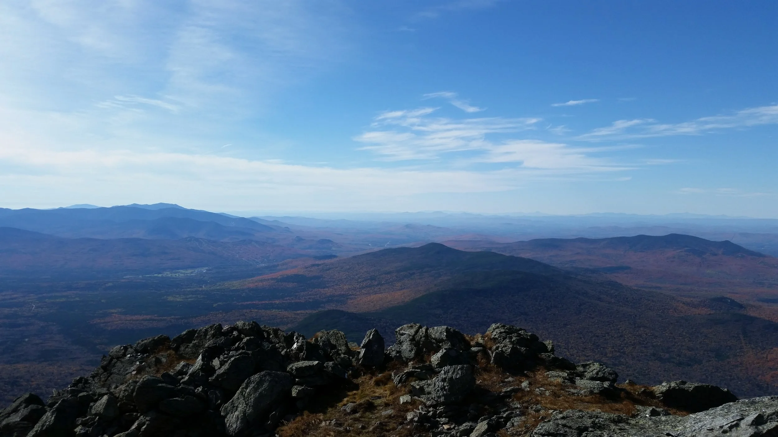 Mountain landscape with rocky foreground, rolling hills, and distant mountain ranges under a blue sky with clouds.