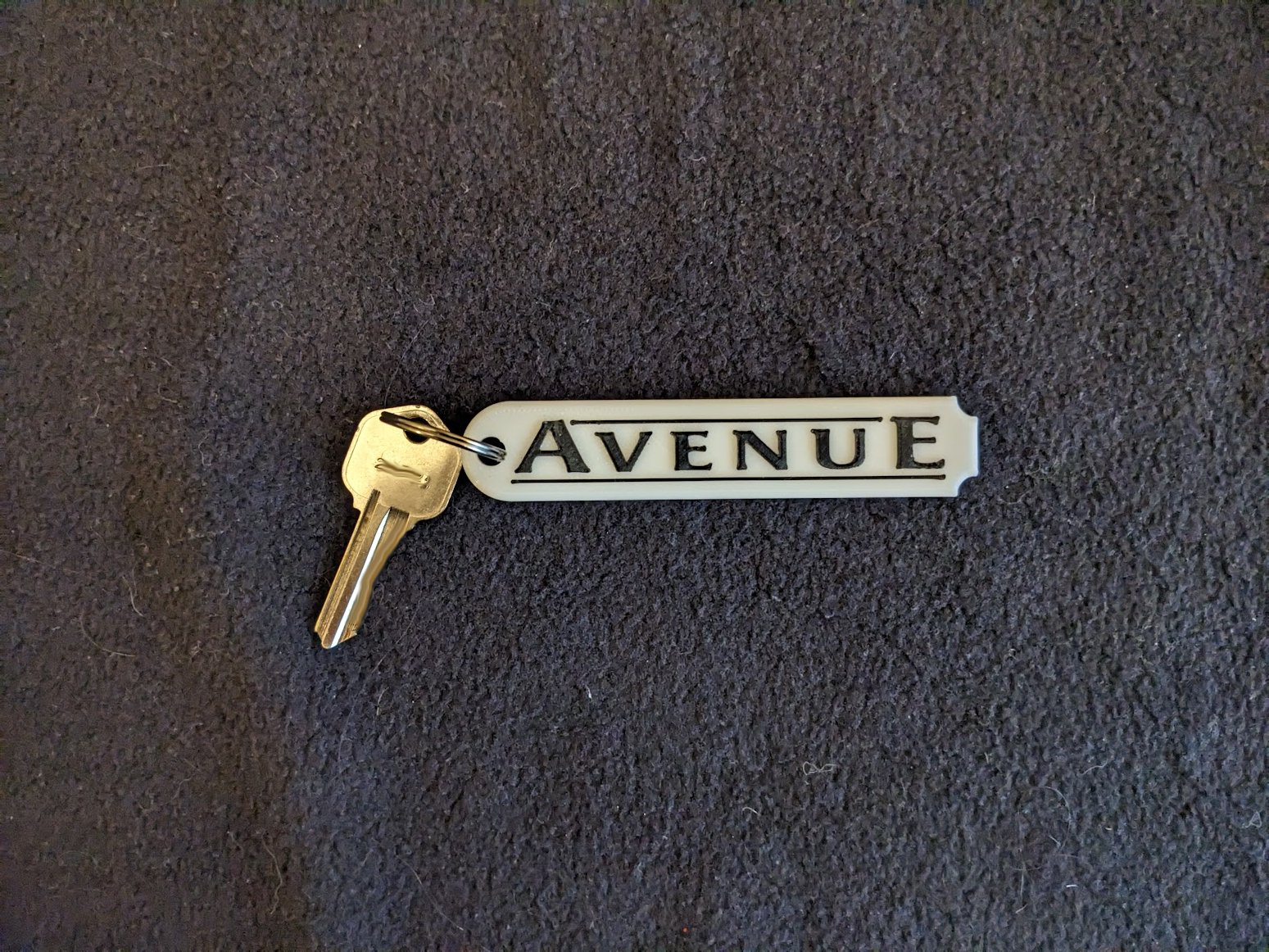 A roughly rectangular white keytag rounded on the left side and scalloped on the right, bearing the Alliance RV model name "Avenue" in black lettering matching the company font and graphics.  Pictured with a gold key attached via a silver keyring