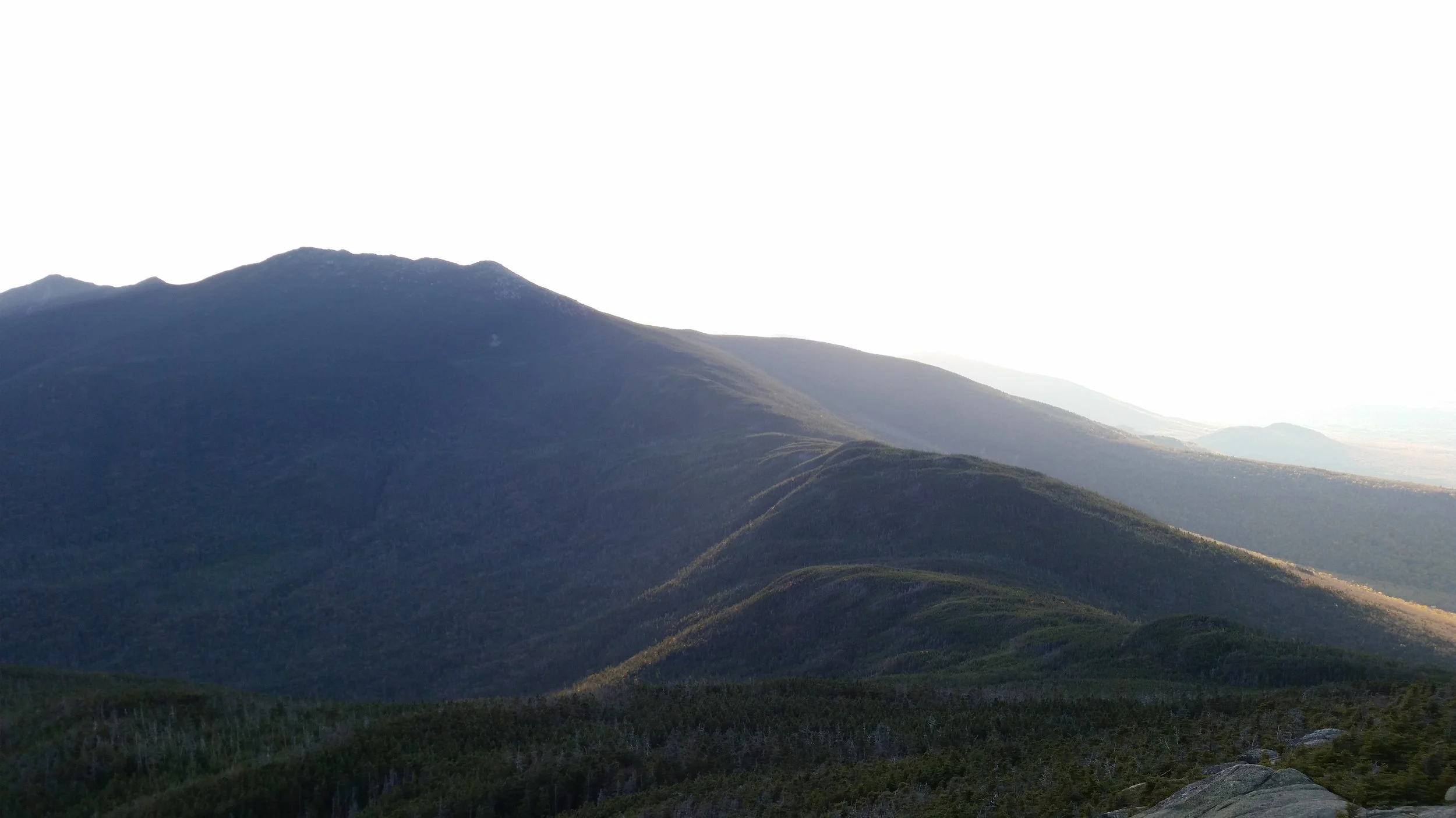 Mountain landscape with rolling hills and a forested foreground, under a bright sky.