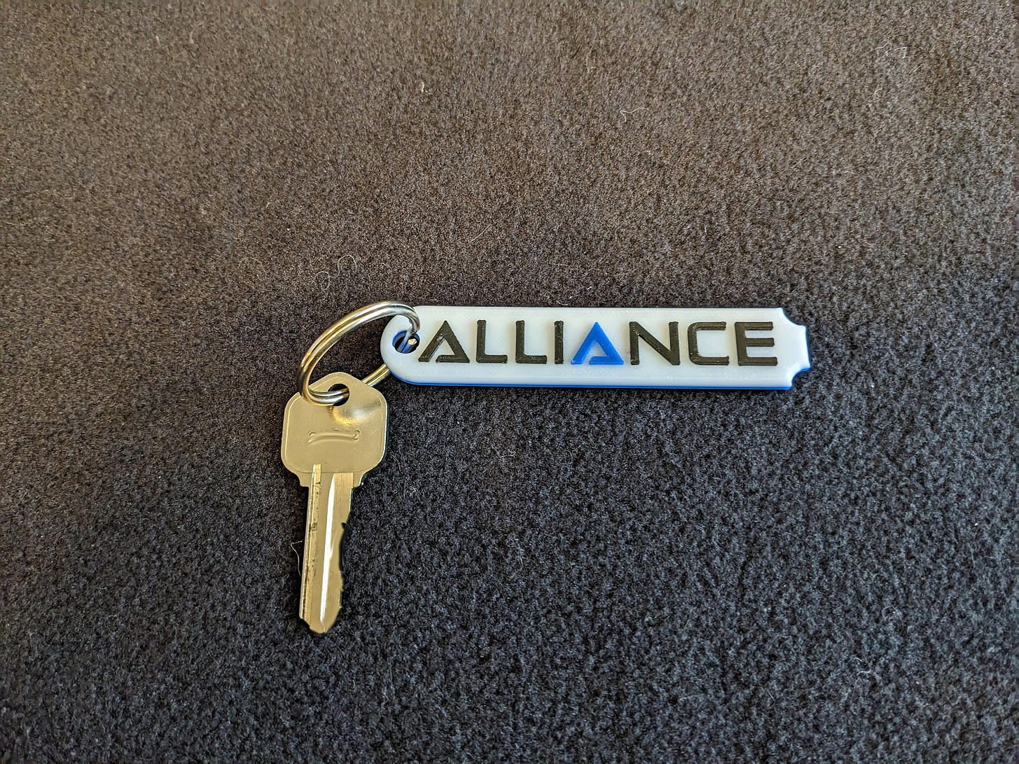 A roughly rectangular white keytag rounded on the left side and scalloped on the right, bearing the RV manufacturer's name "Alliance" in black lettering matching the company font and graphics.  Pictured with a gold key attached via a silver keyring