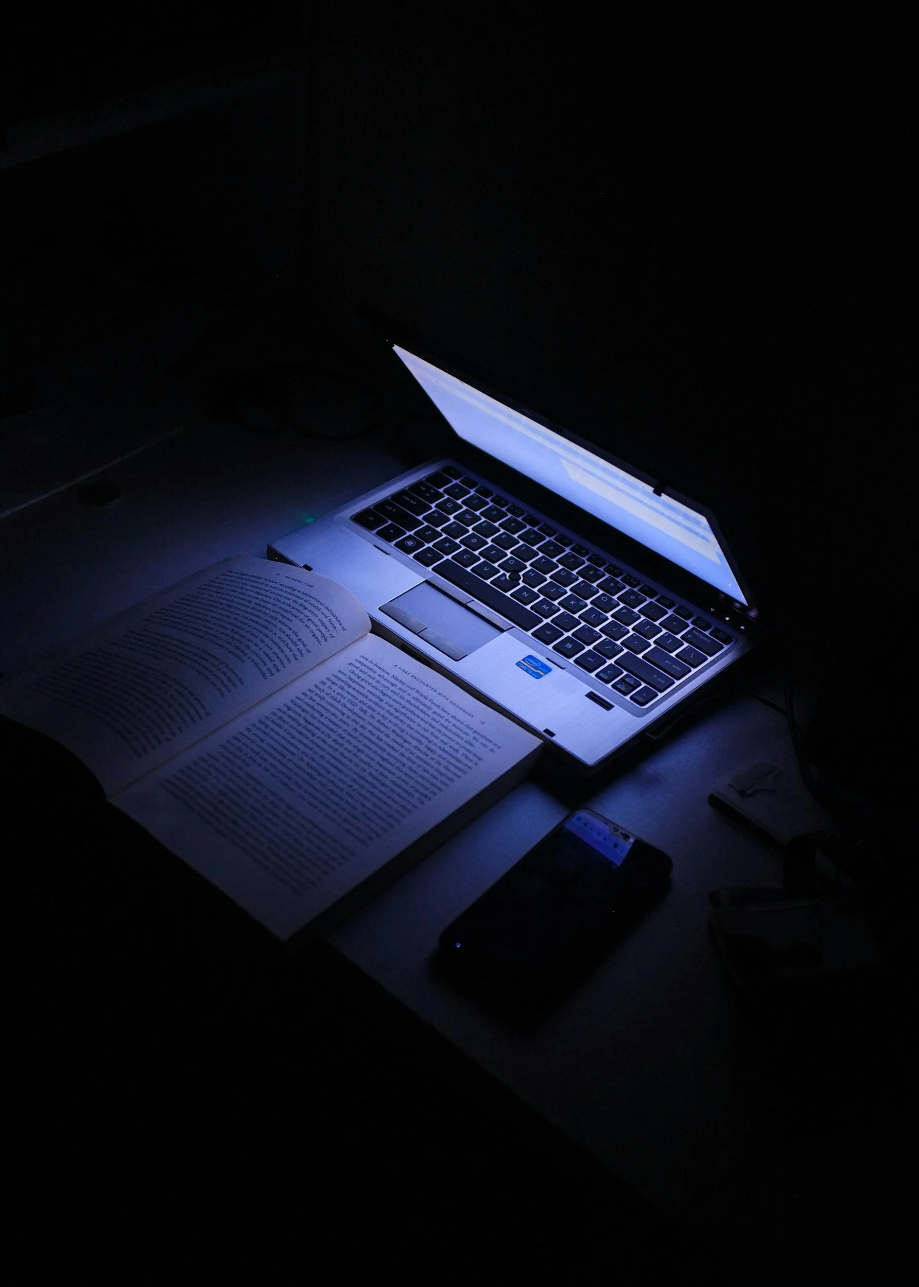 Open book and laptop on a desk in dark surroundings, illuminated by the computer screen.