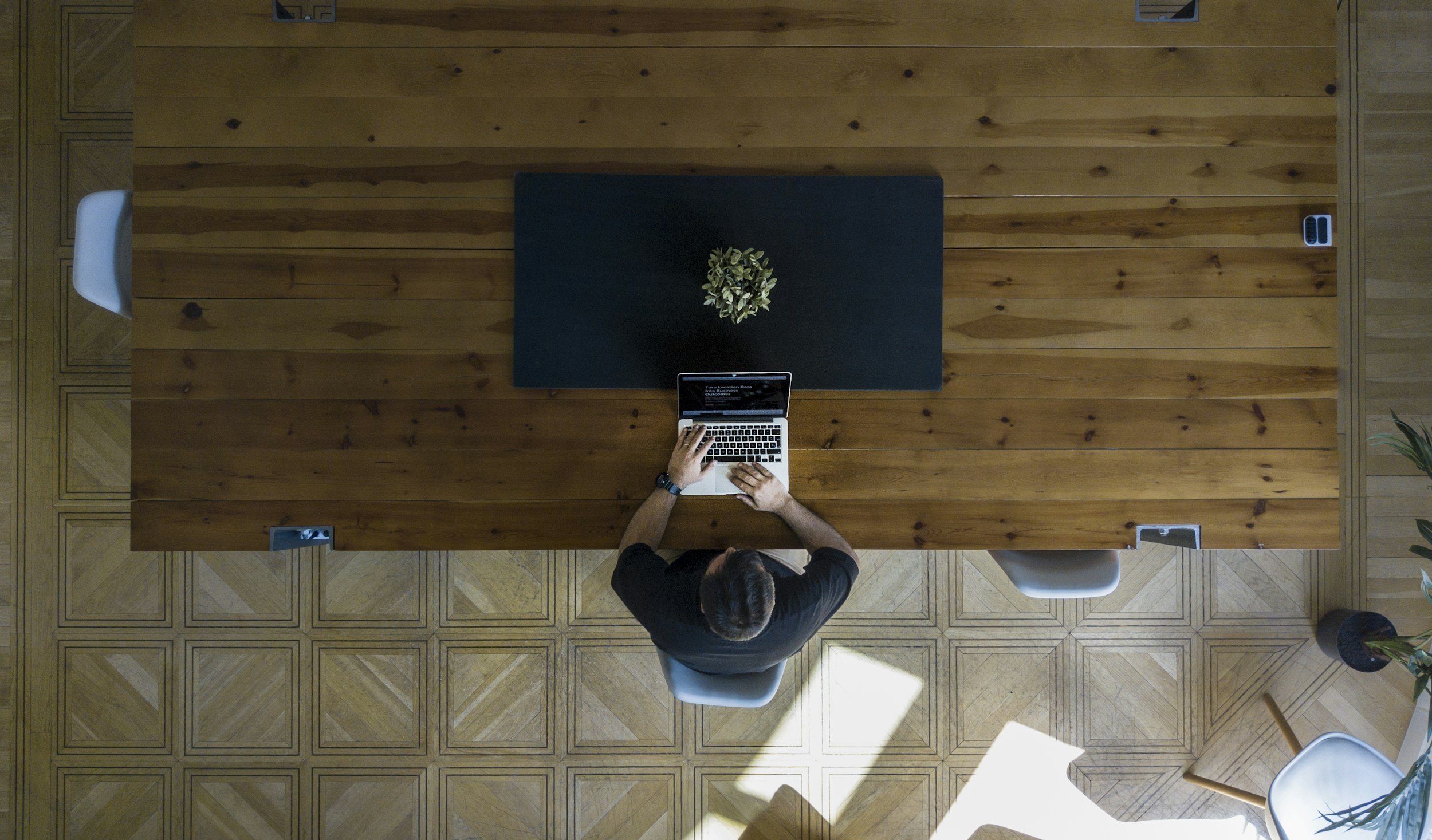 Top-down view of a person sitting at a wooden desk working on a laptop. The person is wearing a black shirt, has dark hair, and a watch on their left wrist. There is a small plant on the desk, and the desk is in a room with patterned wooden flooring and some chairs and plants visible.