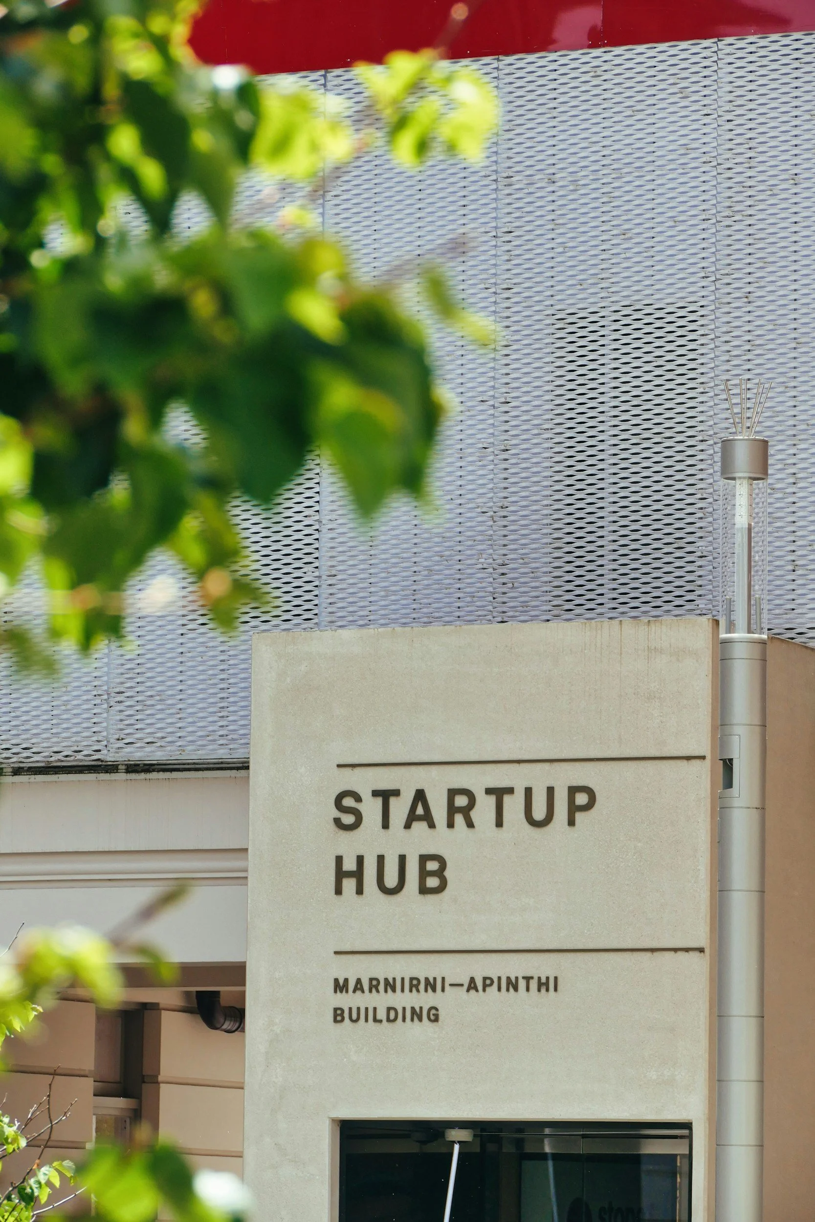 Exterior view of the Startup Hub building, part of the Marnini-APINTHI Building, with a sign and some green leaves in the foreground.