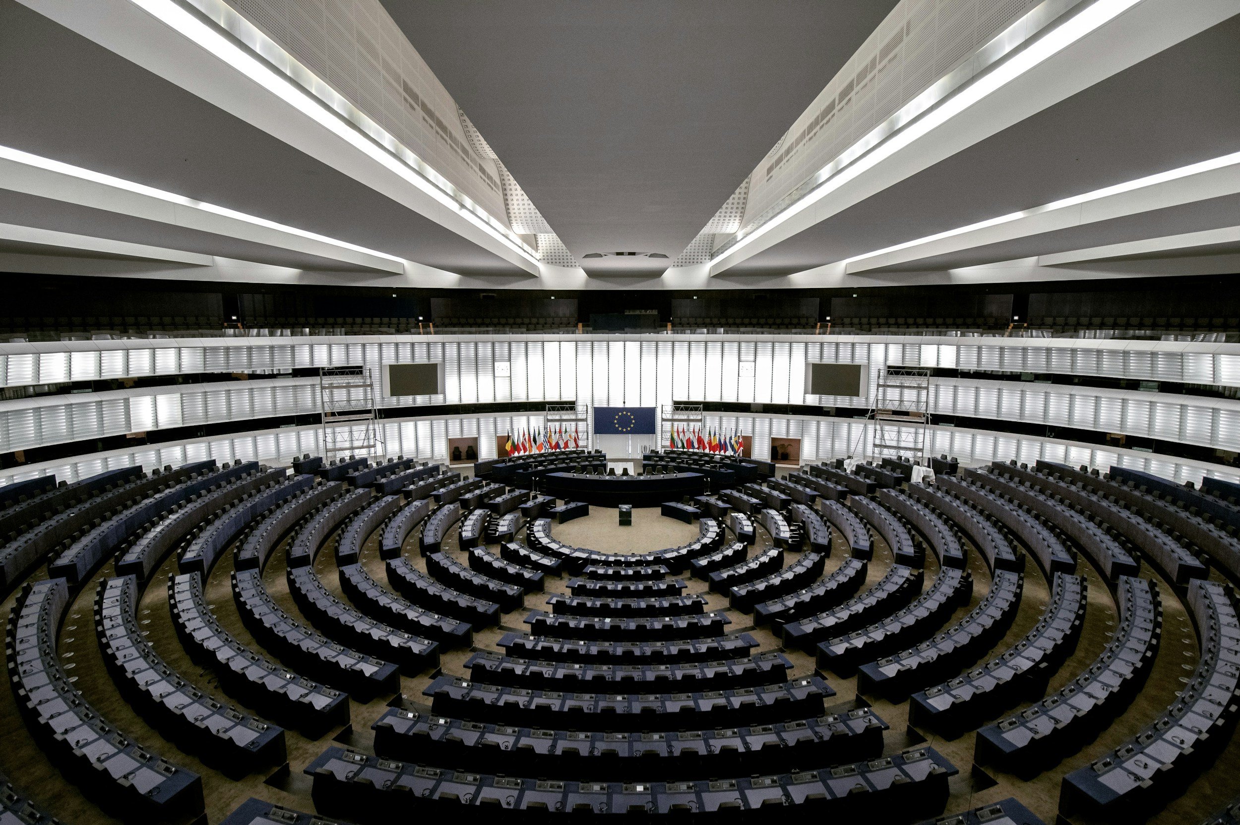 Empty European Union parliamentary chamber with several rows of chairs and desks, a semi-circular seating arrangement, large windows with blinds, and EU flags on a podium, with a European Union flag in the center behind it.