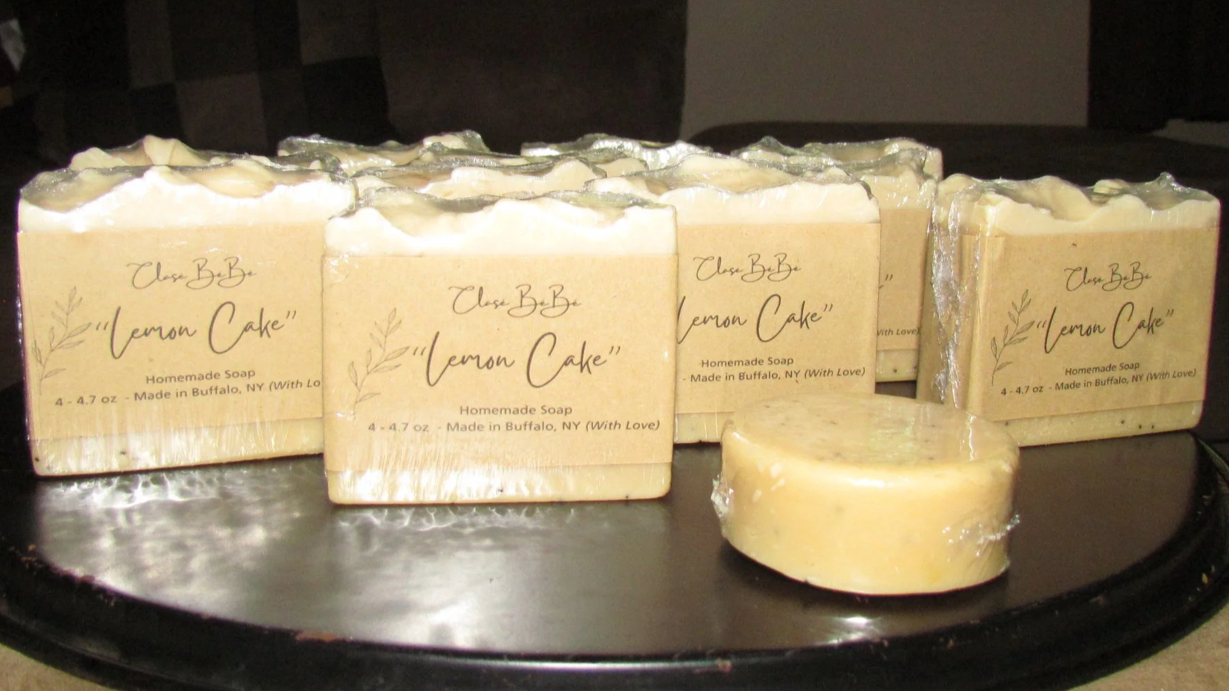 Several handmade soap bars labeled 'Lemon Cake' on a black surface. The soap is yellow with a decorative top and wrapped with beige paper labeling it as homemade soap made in Buffalo, NY.