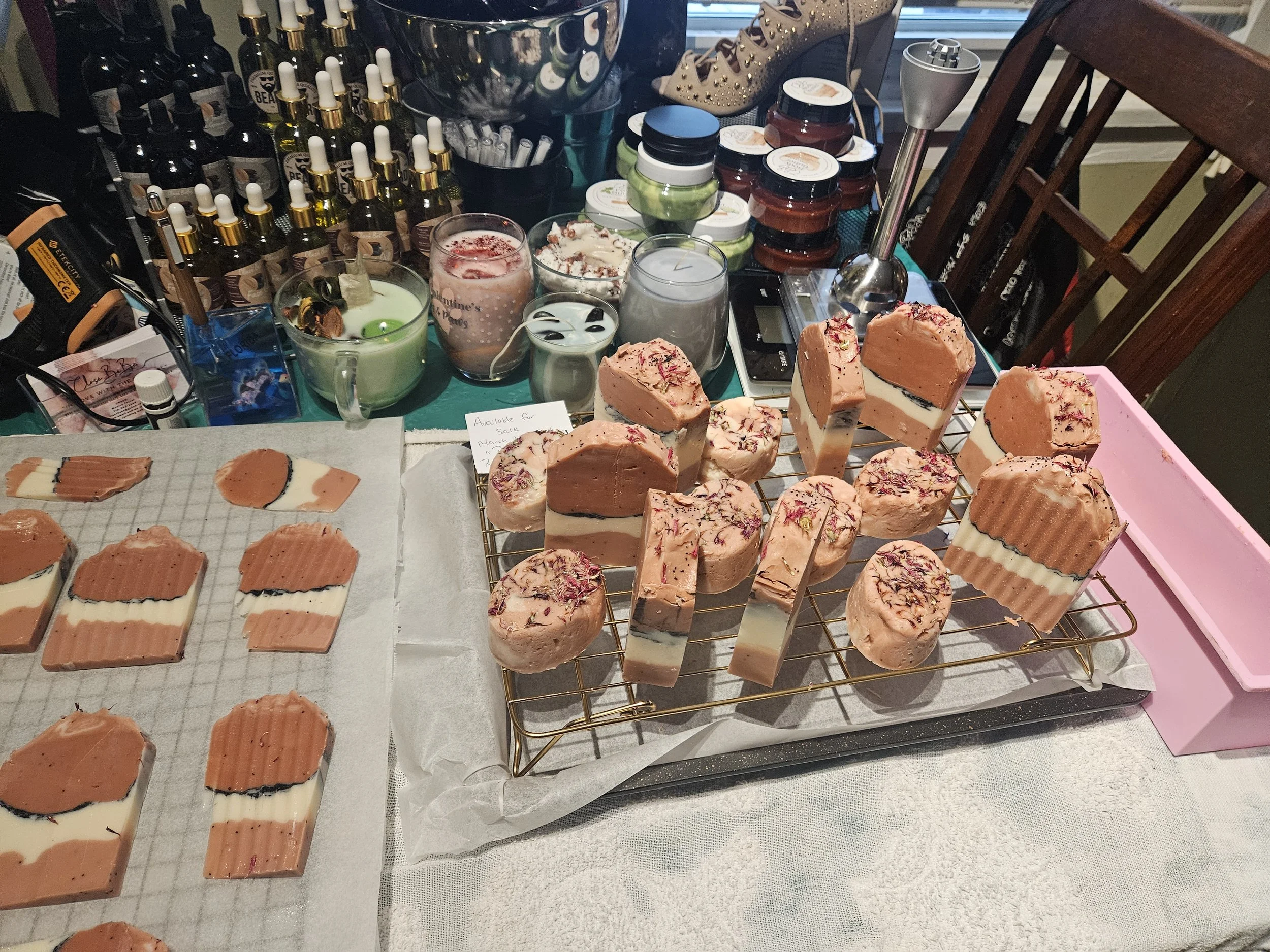 Assorted handmade soaps with pink, white, and black layered designs displayed on a cooling rack, surrounded by various skincare products, candles, and tools on a table.