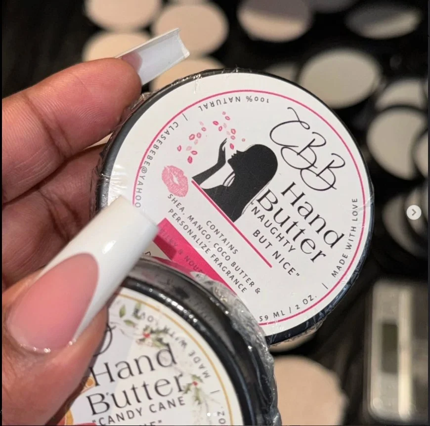 Close-up of a person holding a small container of hand butter with a pink and black label. The label features a silhouette of a woman with long hair, pink lips, and floral design, with text that reads "Hand Butter Naughty But Nice".