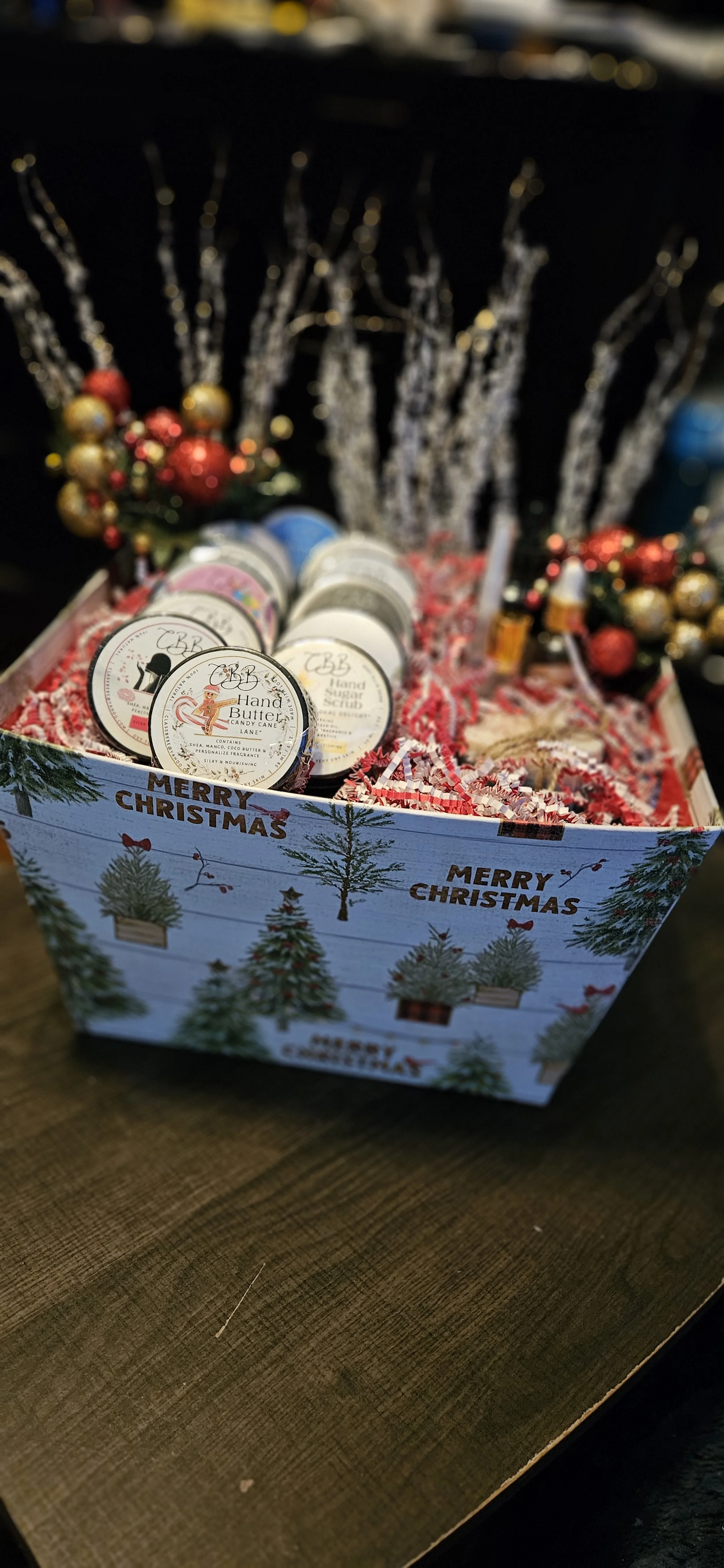 A decorated Christmas gift box containing hand butter scrub jars, with a festive holiday background of pine branches, ornaments, and silver tinsel.
