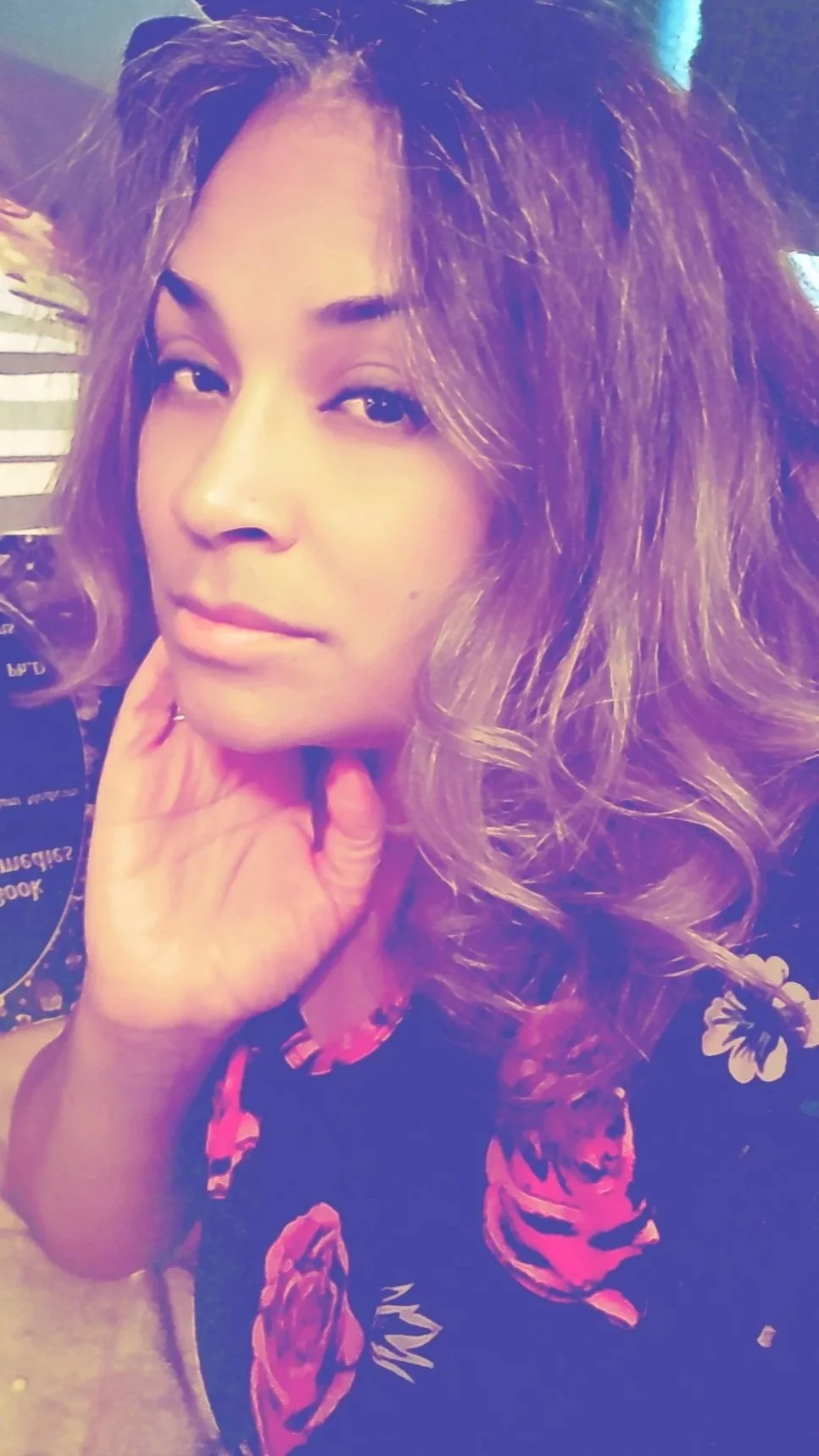 A woman with wavy hair and light makeup resting her chin on her hand, wearing a floral dress with pink and red flowers.