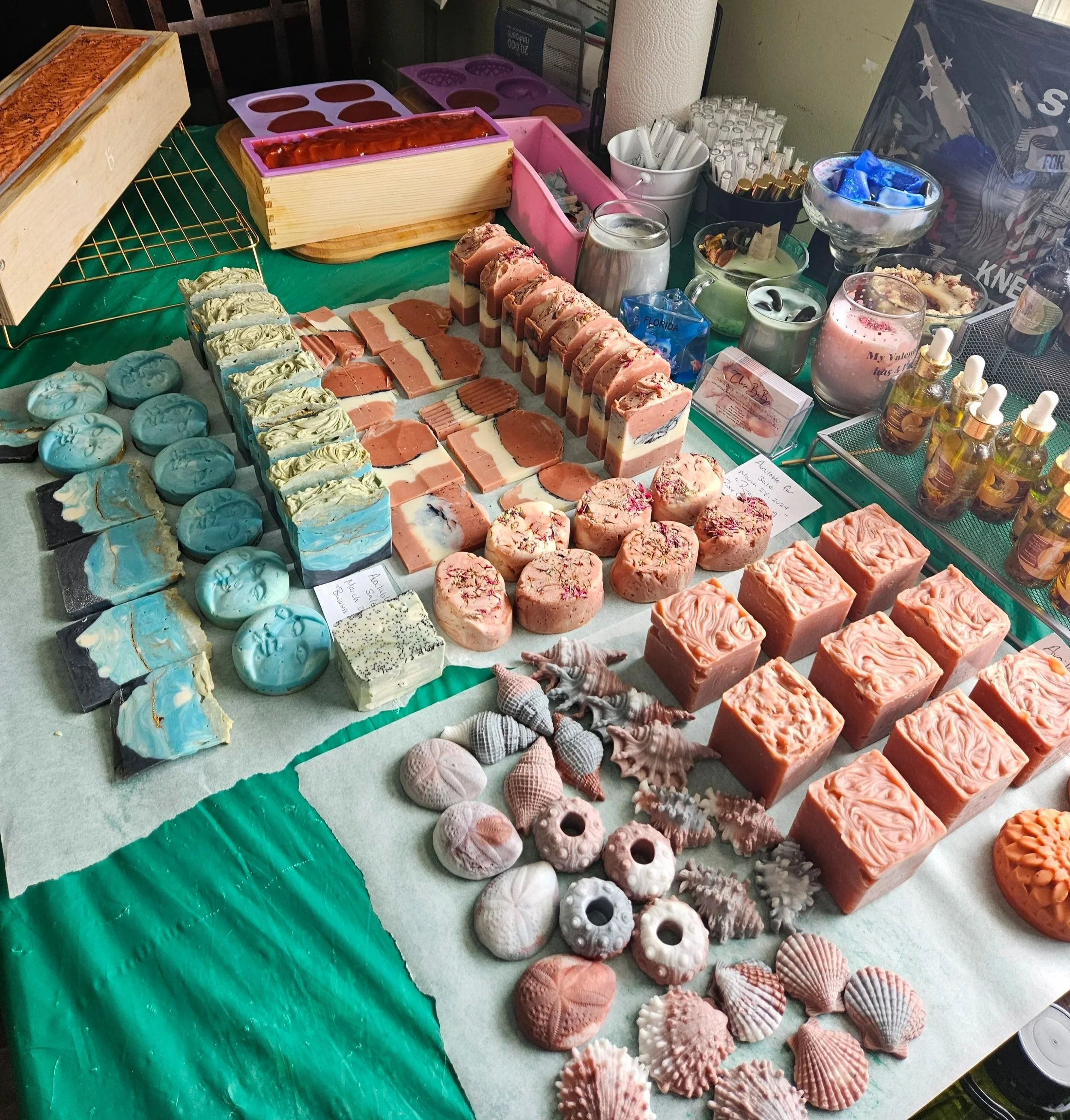 A table with various handmade soaps, including swirling blue, orange, and pink soap bars, some with decorative elements, and palette-shaped soap pieces. The table also has decorative shells and other sea-themed items.