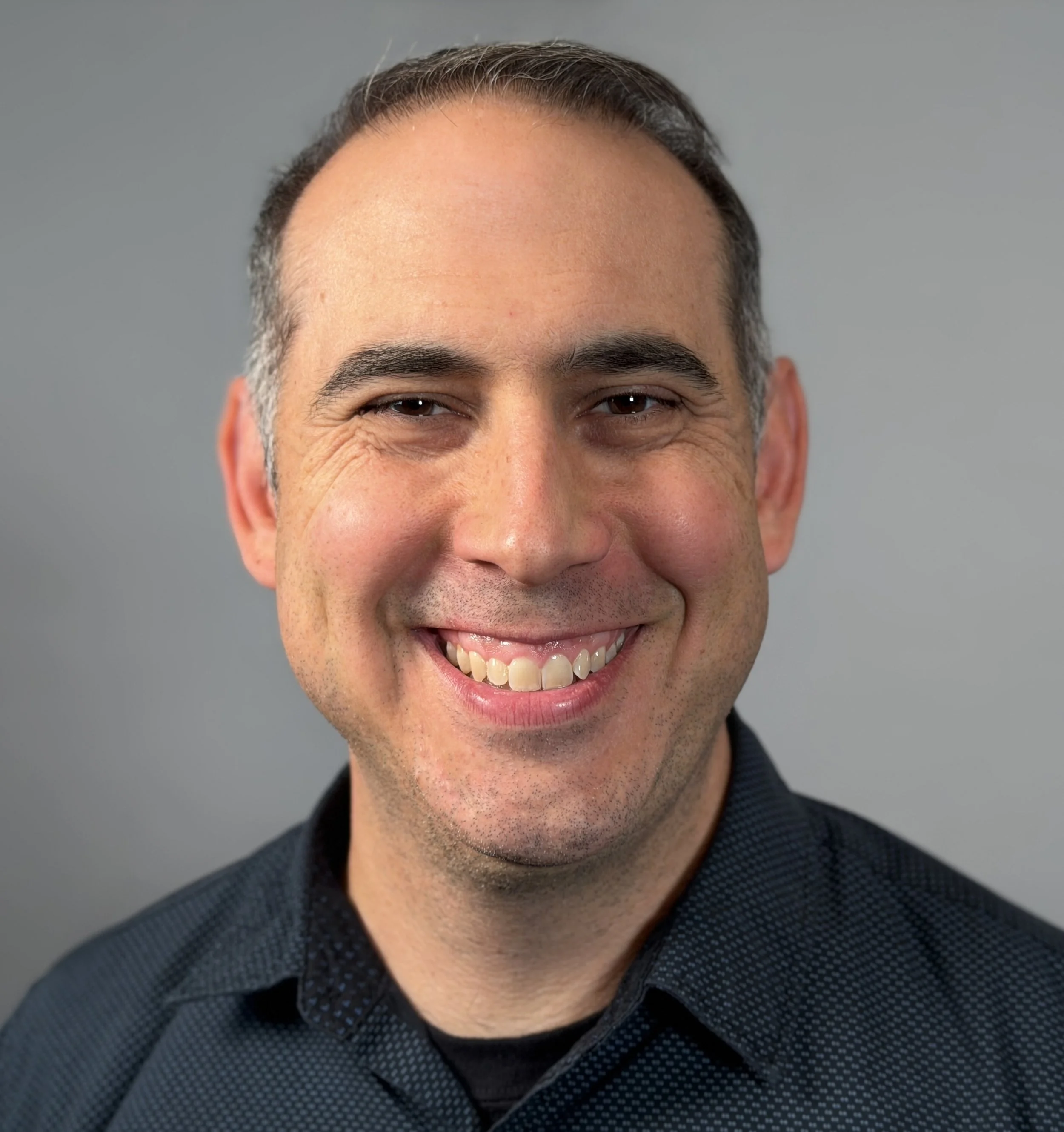 Close-up of a smiling man with dark hair, wearing a dark button-up shirt.