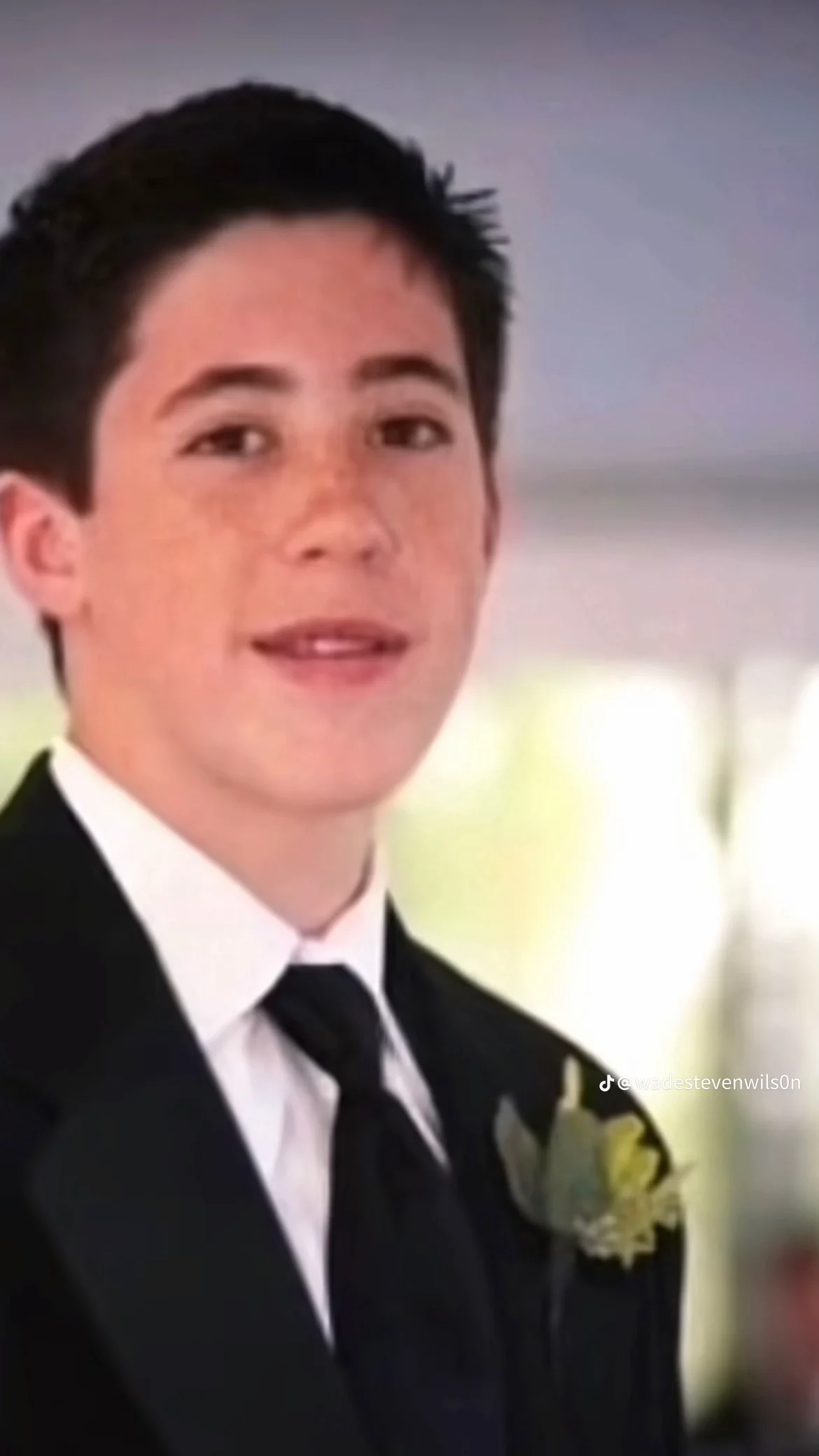 Young man dressed in a suit with a white shirt and black tie, wearing a boutonniere, smiling indoors.