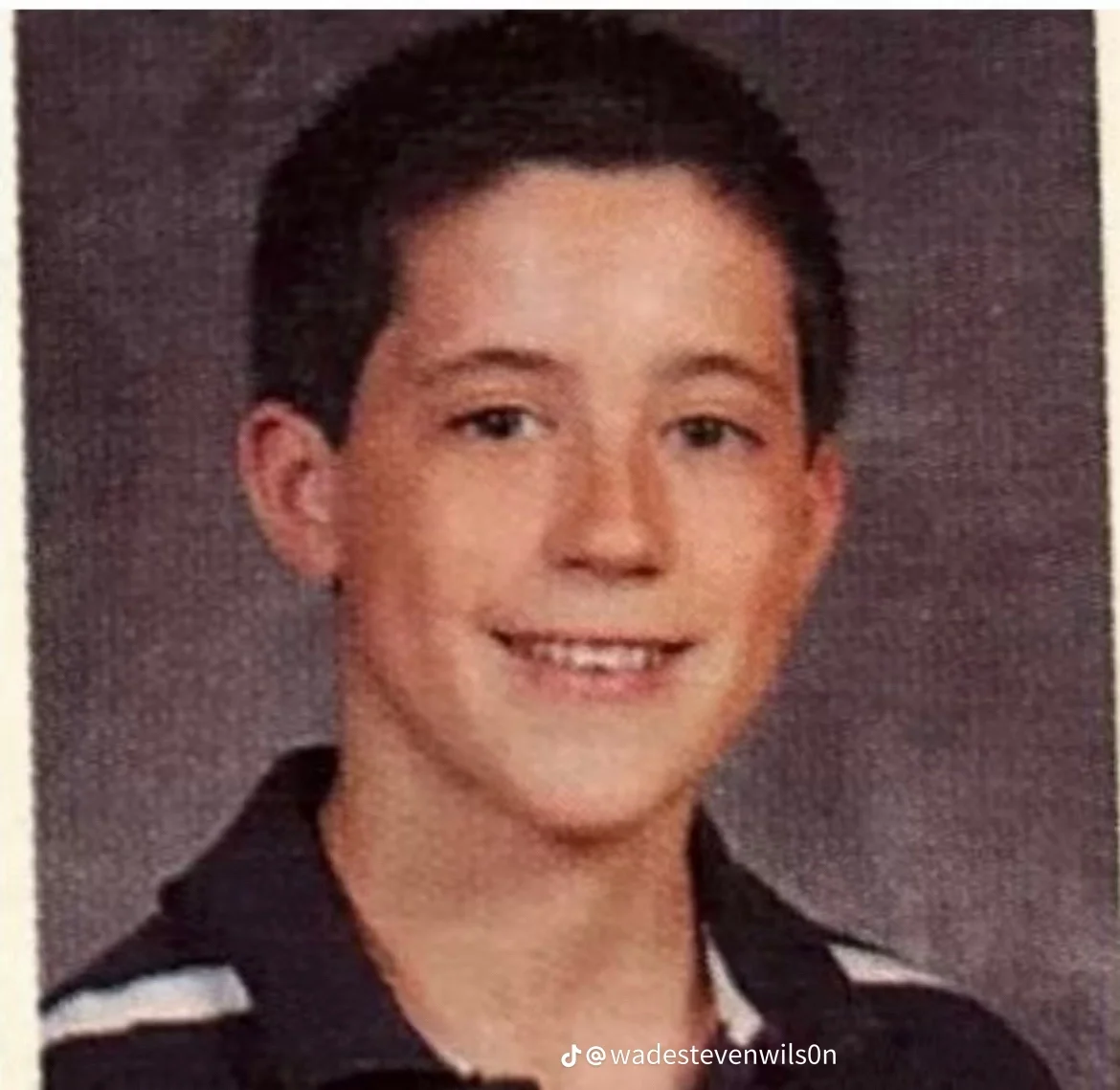 A young boy with short dark hair, smiling at the camera, wearing a dark shirt with white stripes on the shoulder, in front of a dark background.