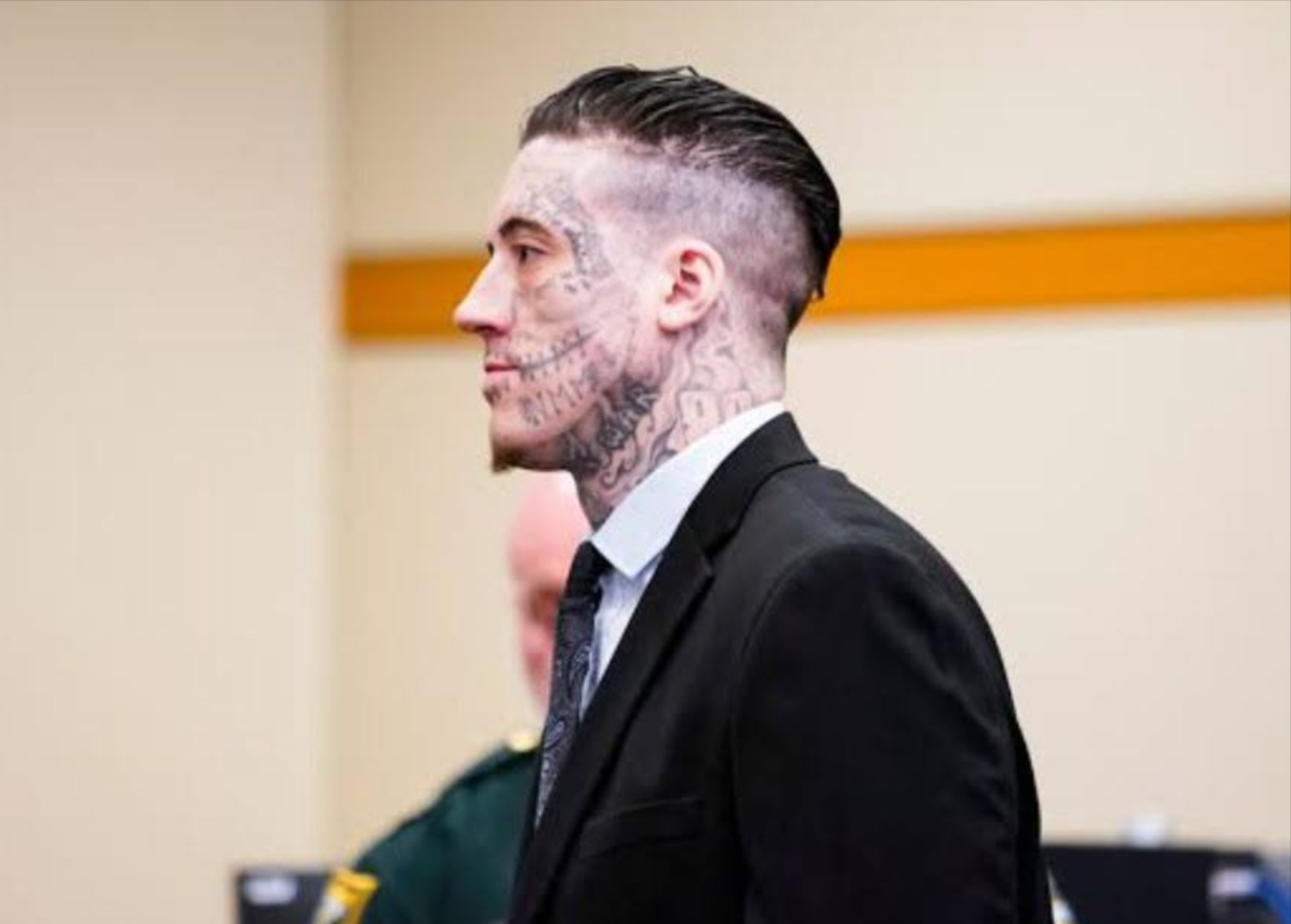 A man with tattoos on a skull and face, dressed in a black suit and tie, is standing in a courtroom.