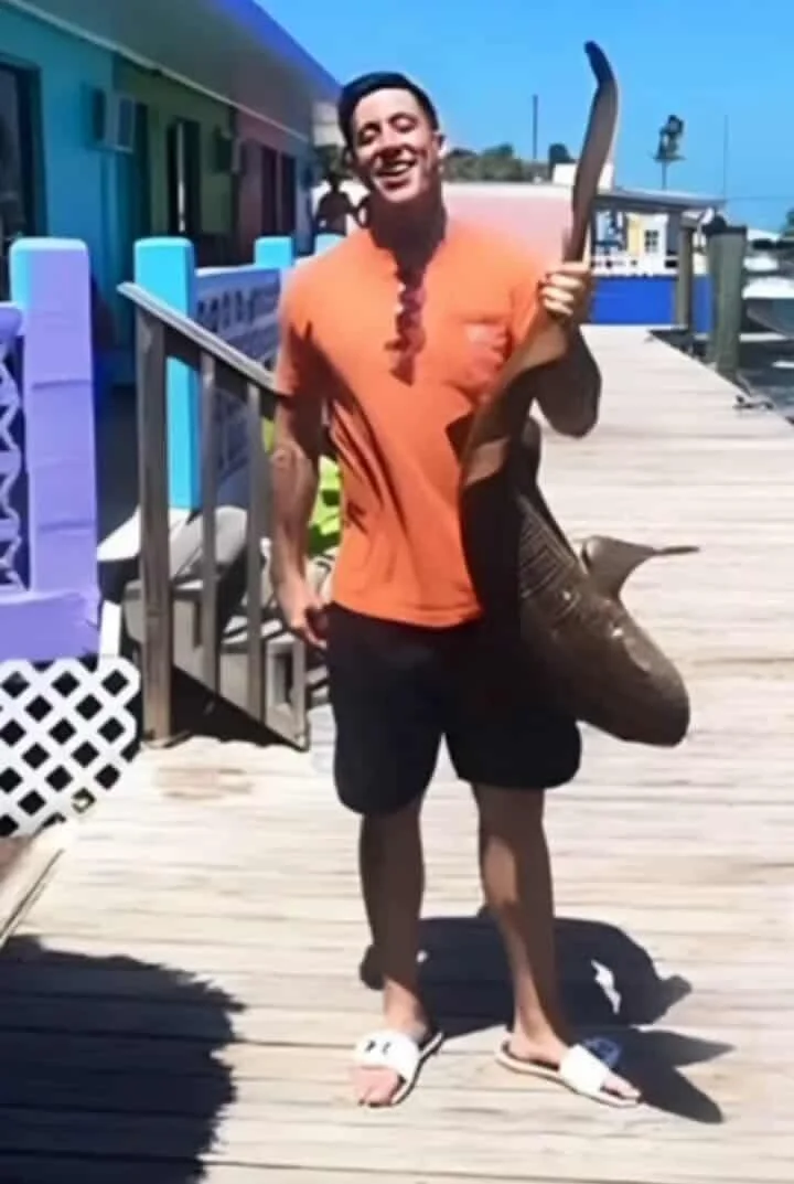 A man standing on a wooden dock near colorful painted buildings, holding a large fish, smiling, dressed in an orange T-shirt, black shorts, and white slides, under a bright blue sky.