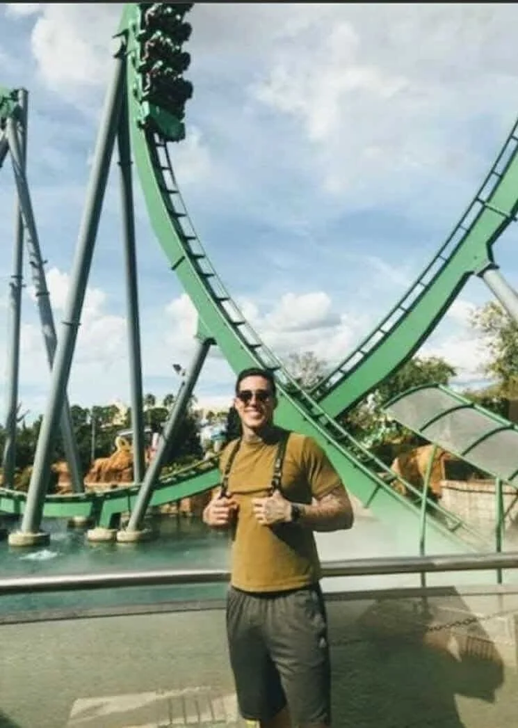 Man standing in front of a green roller coaster at an amusement park, wearing sunglasses, a brown shirt, and gray shorts.