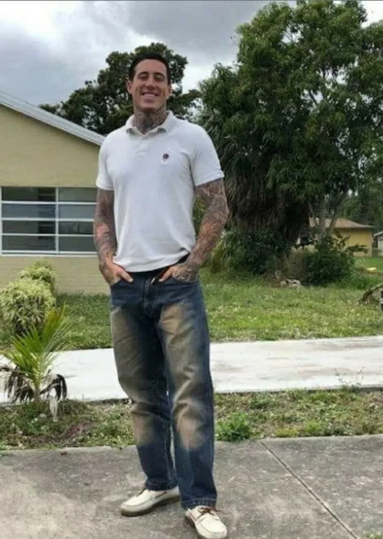 A smiling man with tattoos on his arms, wearing a white polo shirt, jeans, and white shoes, standing outside on a sidewalk with trees, plants, and a house in the background during cloudy weather.