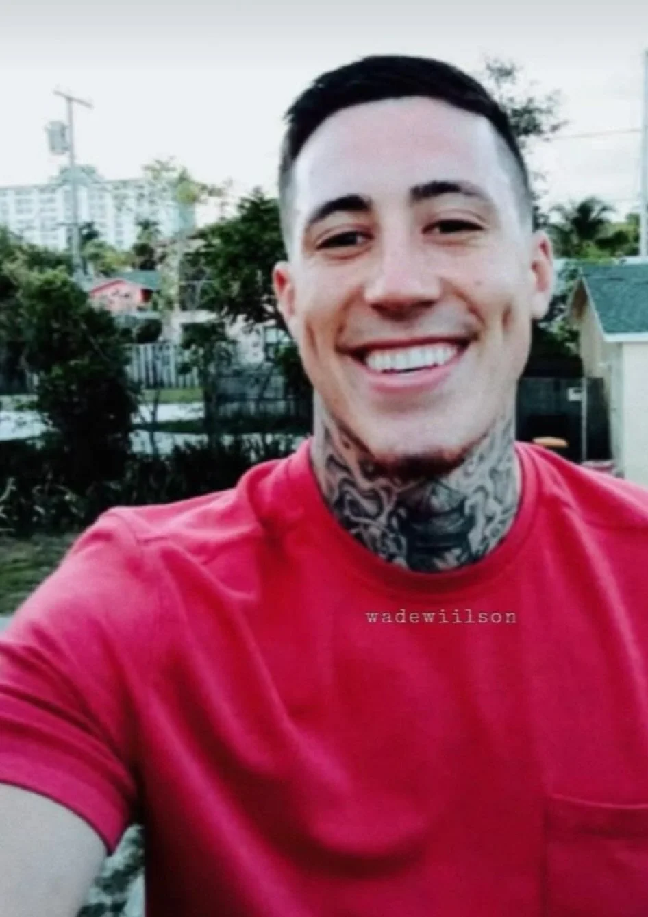Smiling young man with short dark hair, visible tattoos on neck, wearing a red t-shirt, outdoors in a backyard with trees and houses in the background.
