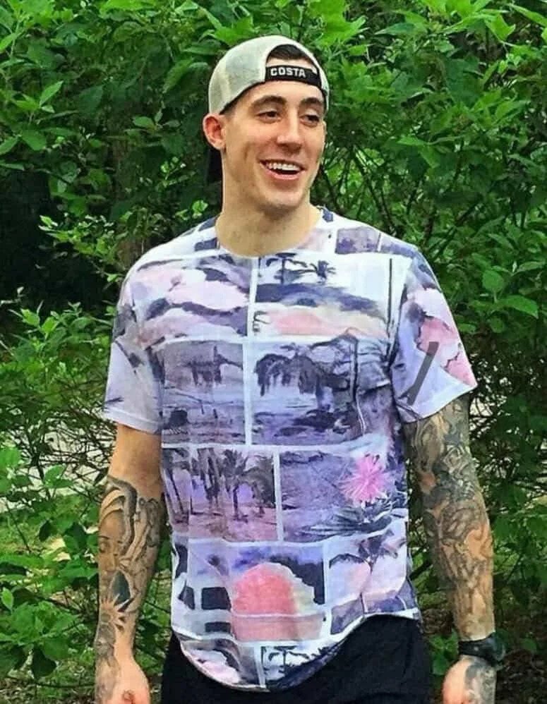 A young man with tattoos on both arms, wearing a casual tropical-themed T-shirt featuring palm trees and sunsets, a gray baseball cap worn backward, and a black wristwatch, standing outdoors in front of green foliage, smiling and looking to his left.