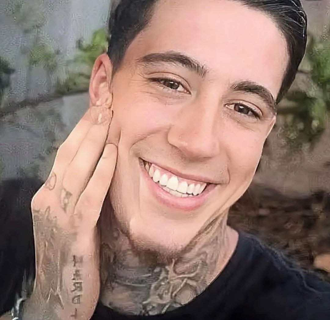 Close-up of a smiling young man with tattoos on hand and neck, touching his face with one hand, outdoor background.