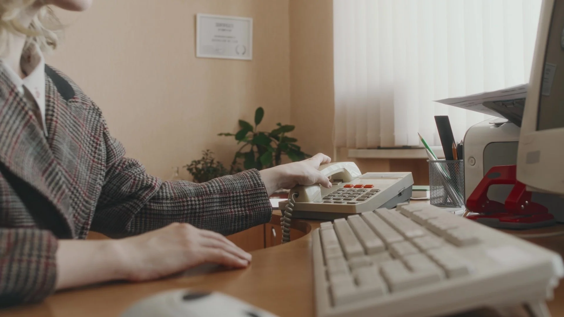 Person in plaid jacket making a phone call at a desk with a computer, keyboard, and office supplies, in an office setting.