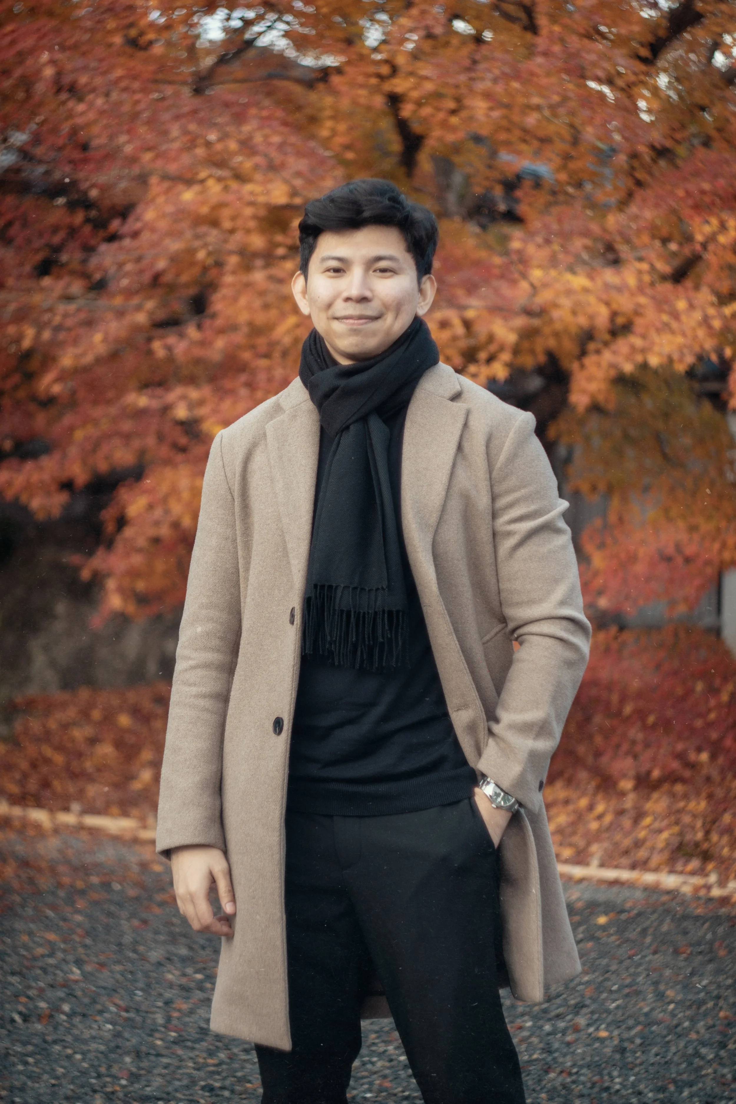A young man in a beige coat and black scarf posing outdoors in front of orange fall foliage.