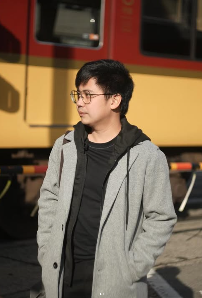 A young man with glasses and black hair stands outdoors near a train, wearing a gray coat over a black hoodie and T-shirt, with hands in coat pockets, looking to the side.