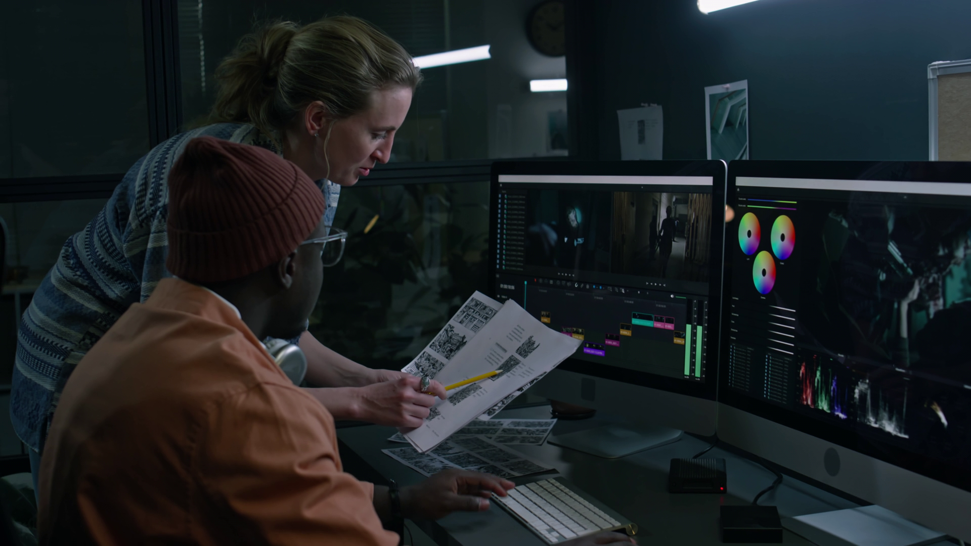 Two women working on video editing at a desk with dual monitors, one displaying video editing software and color grading tools, the other showing a video scene.